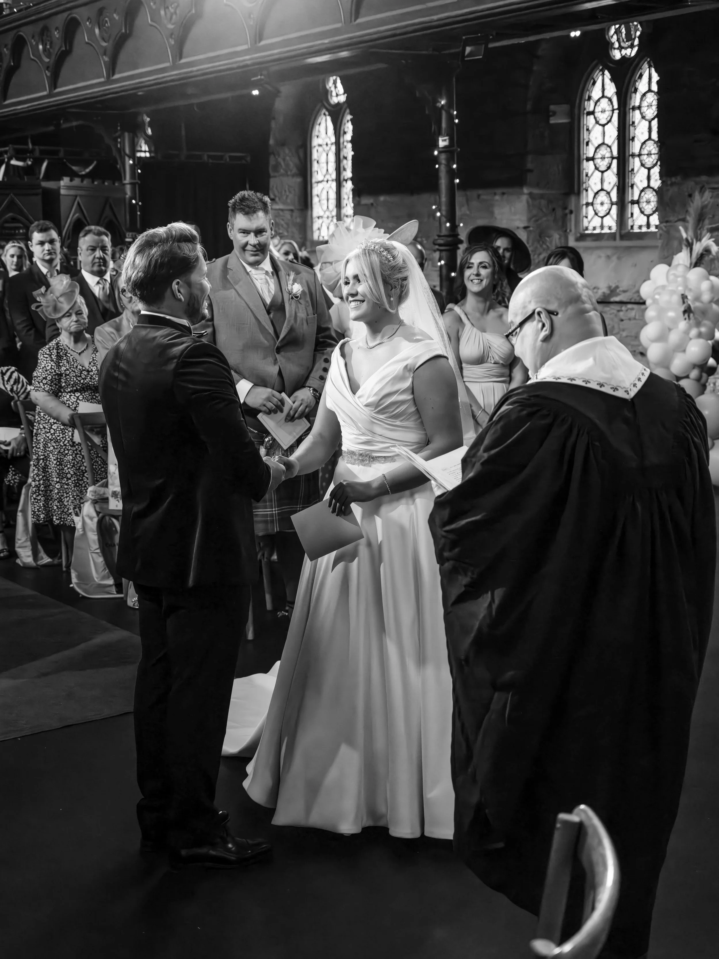 A black and white photo of a wedding ceremony in a church with stained glass windows. The bride and groom are holding hands, smiling at each other, standing before an officiant. Guests are seated and standing around, watching the couple.