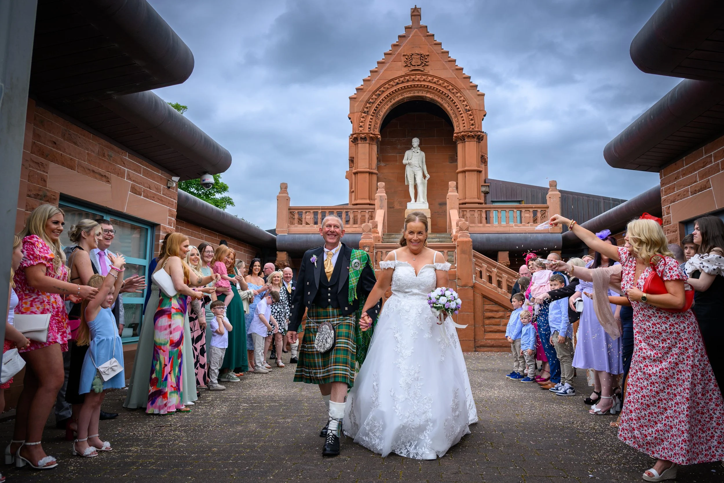 A newly married couple walking hand in hand through a crowd of guests on a wedding day outdoors, with a historic brick building and a statue in the background.