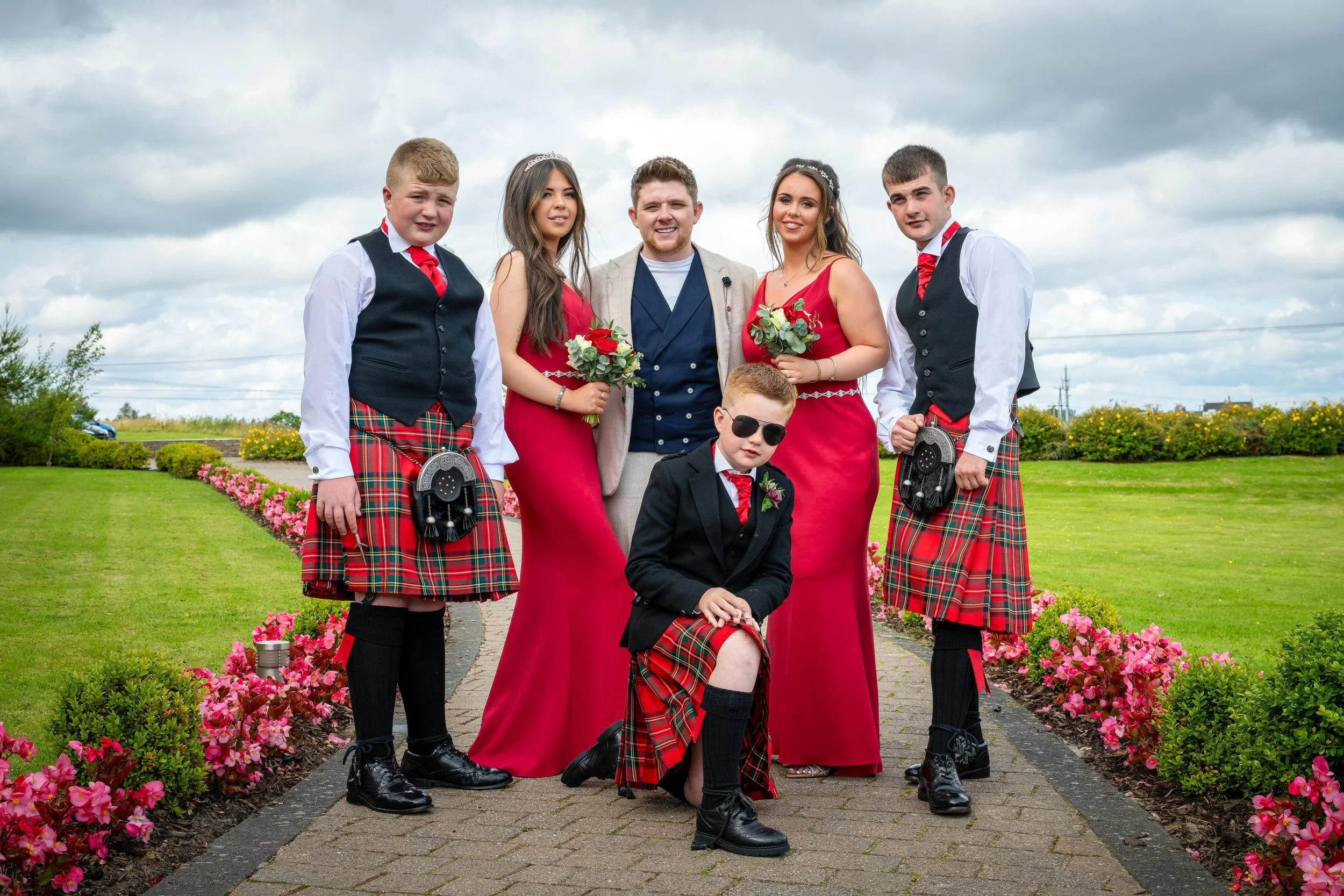 Group of people dressed in wedding attire, including bridesmaids in red dresses, groomsmen in kilts, and a young boy in a kilt, standing on a pathway surrounded by colorful flowers.