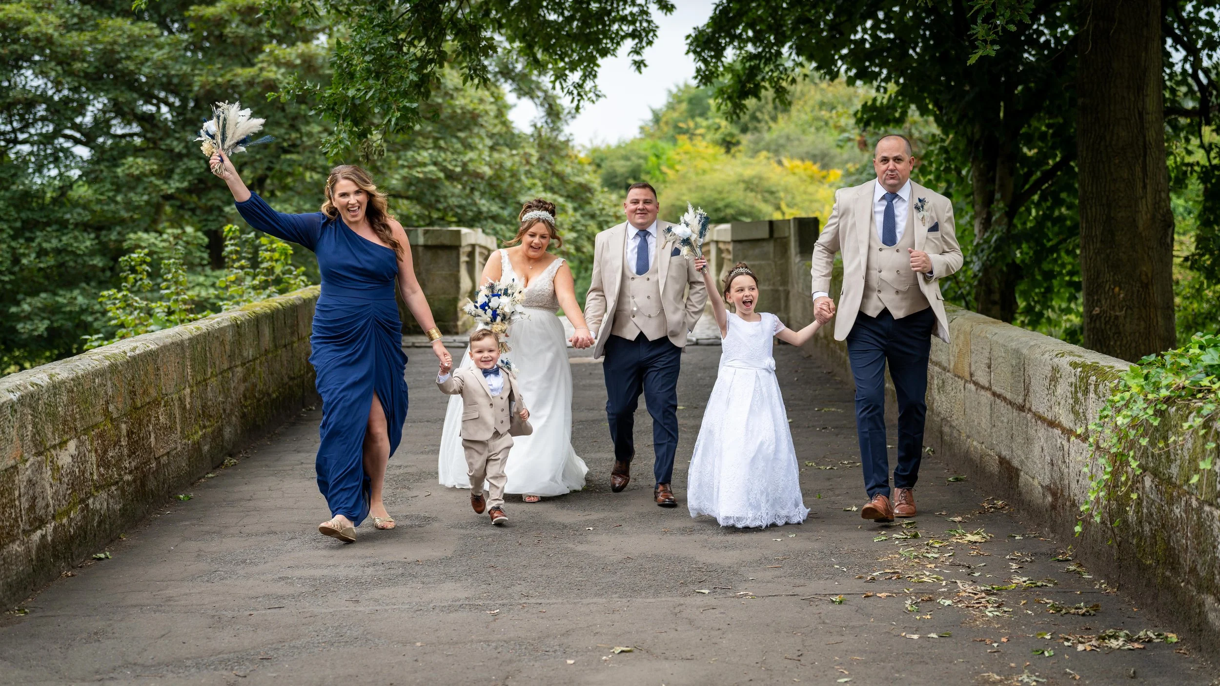 A wedding party walking across a stone bridge over a creek, with a woman in a blue dress, a bride in a white gown, a groom in a beige suit, a young girl in a white dress, and a young boy in a beige suit, holding bouquets and smiling.