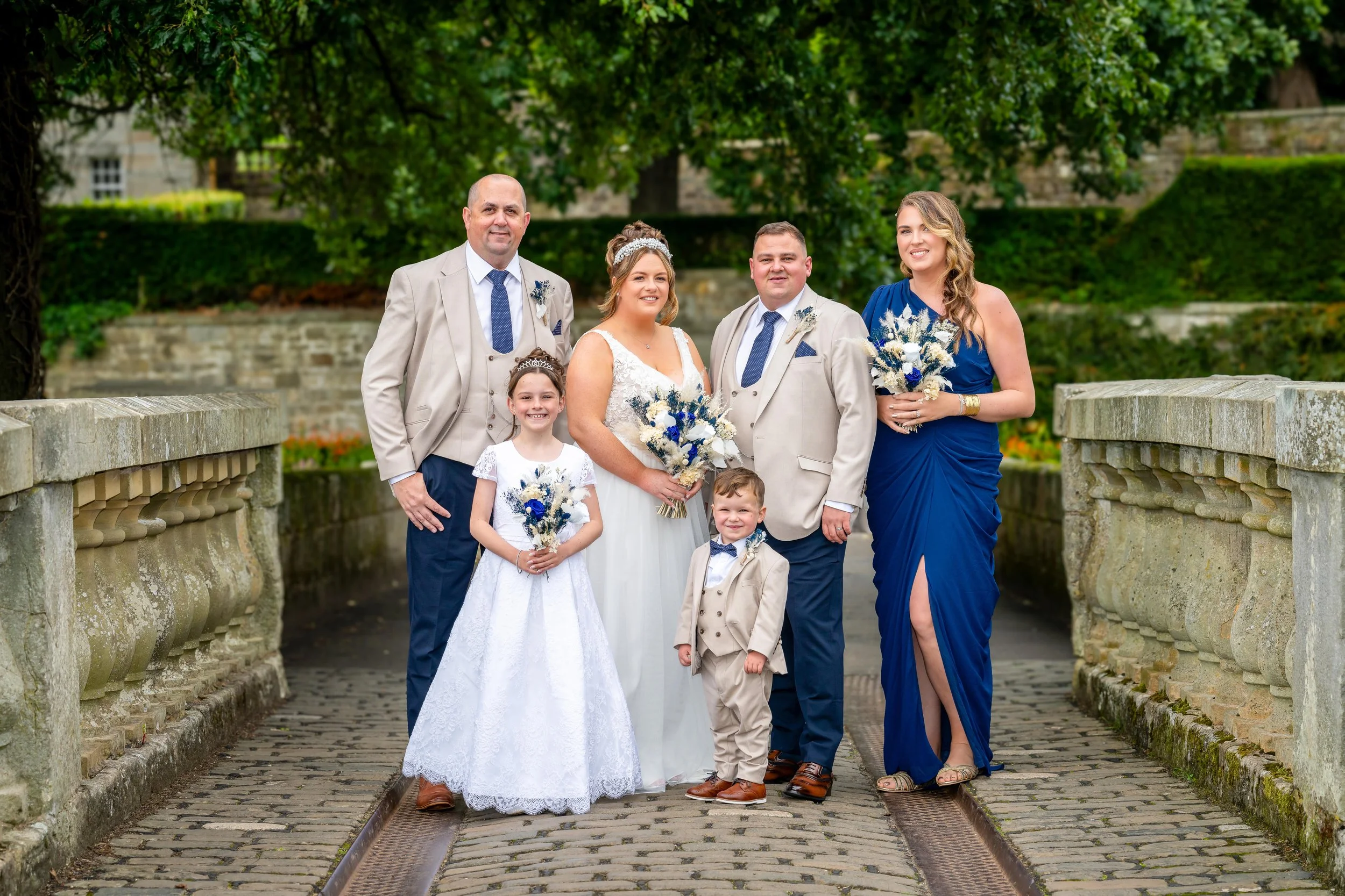 A wedding party standing on a stone bridge outdoors, including a bride, groom, two children, and three adults, all dressed in formal attire with flowers.