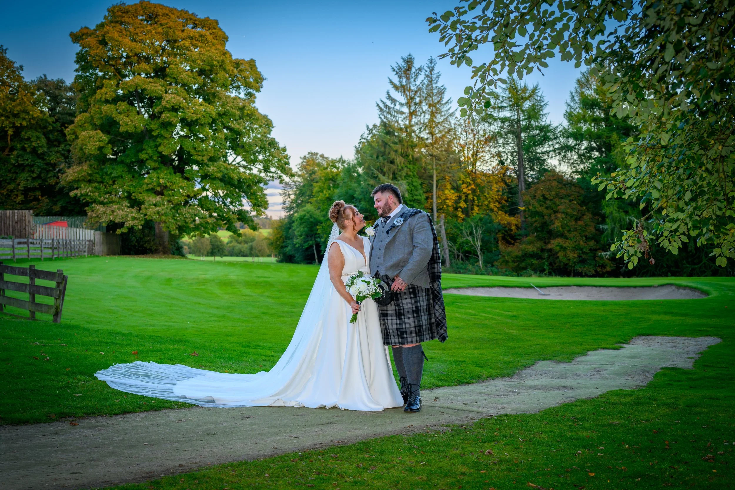 A bride and groom standing on a dirt path in a lush green park during sunset, sharing a moment of affection. The bride is wearing a long white wedding gown and holding a bouquet, while the groom is dressed in traditional Scottish attire, including a 