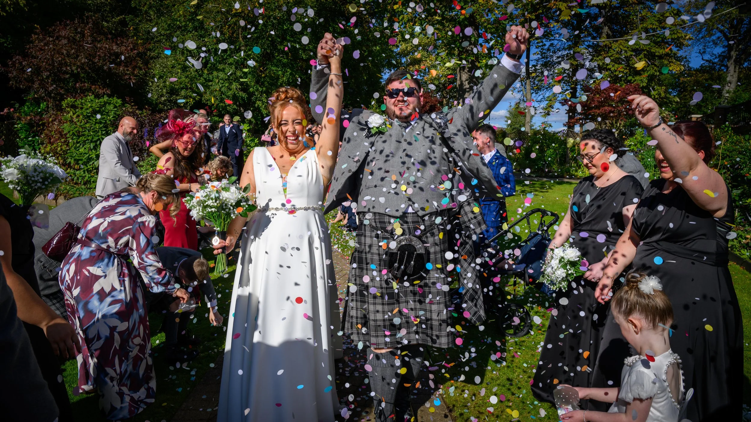 Couple celebrating their wedding in a garden with confetti, surrounded by friends and family.
