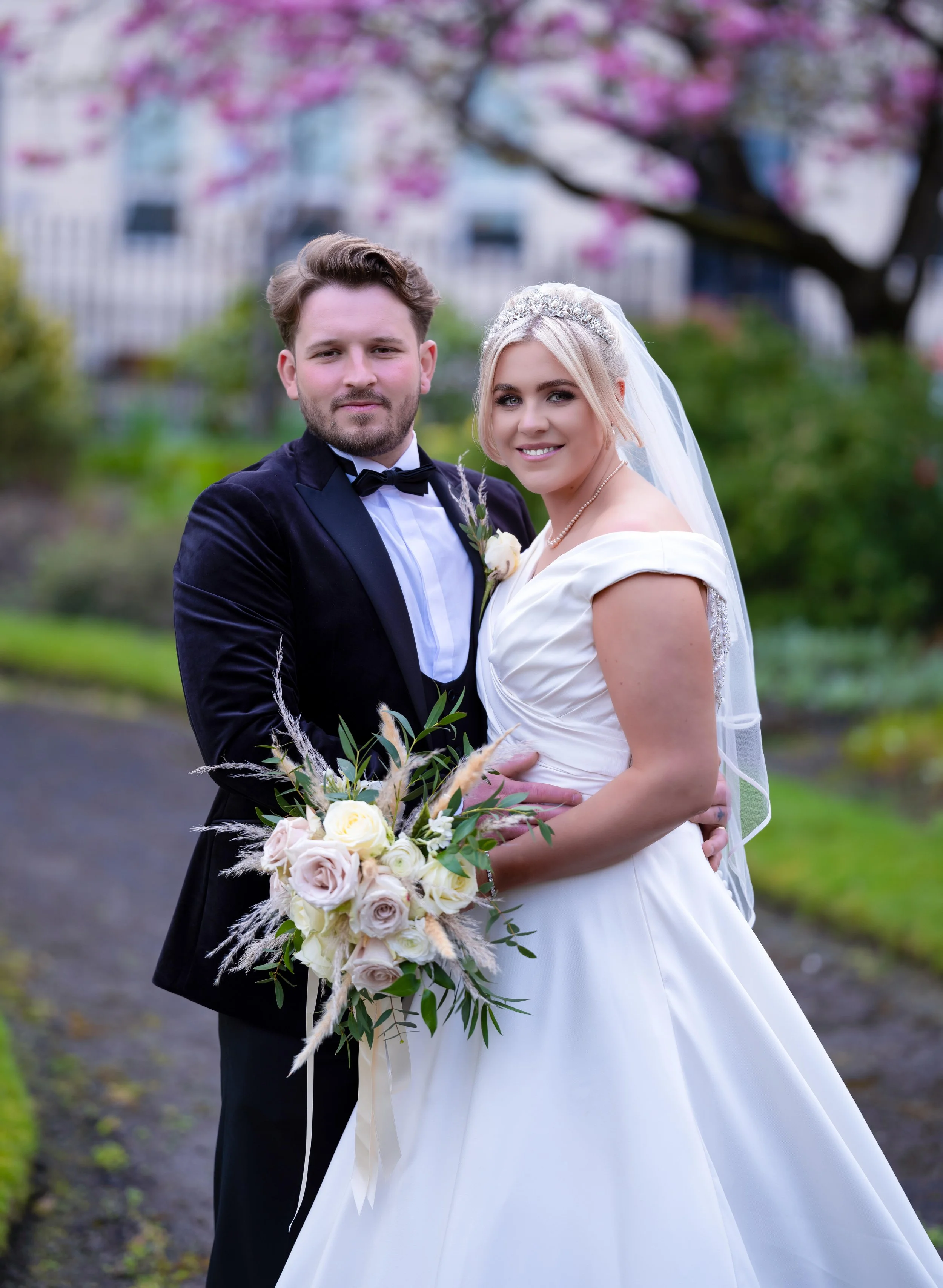 A newlywed couple standing outdoors during spring, with pink blossoming trees in the background. The groom is wearing a black tuxedo with a bow tie, and the bride is in a white wedding gown with a veil, holding a bouquet of white and pale pink roses 