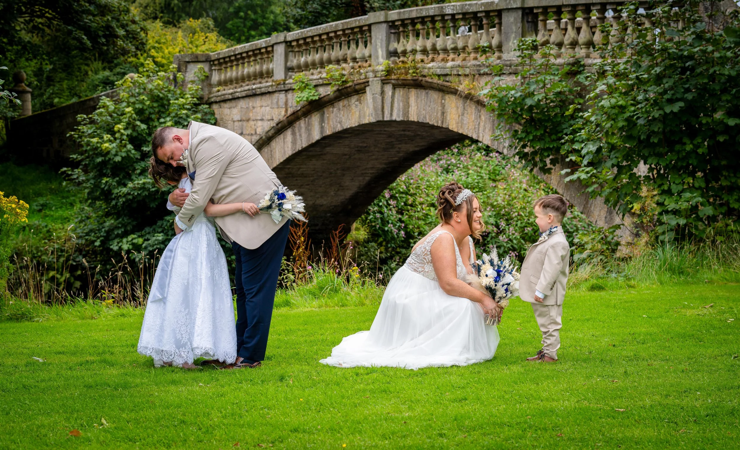 A wedding scene outdoors with two women in white wedding dresses and two boys in beige suits, under a stone bridge with greenery and flowers.
