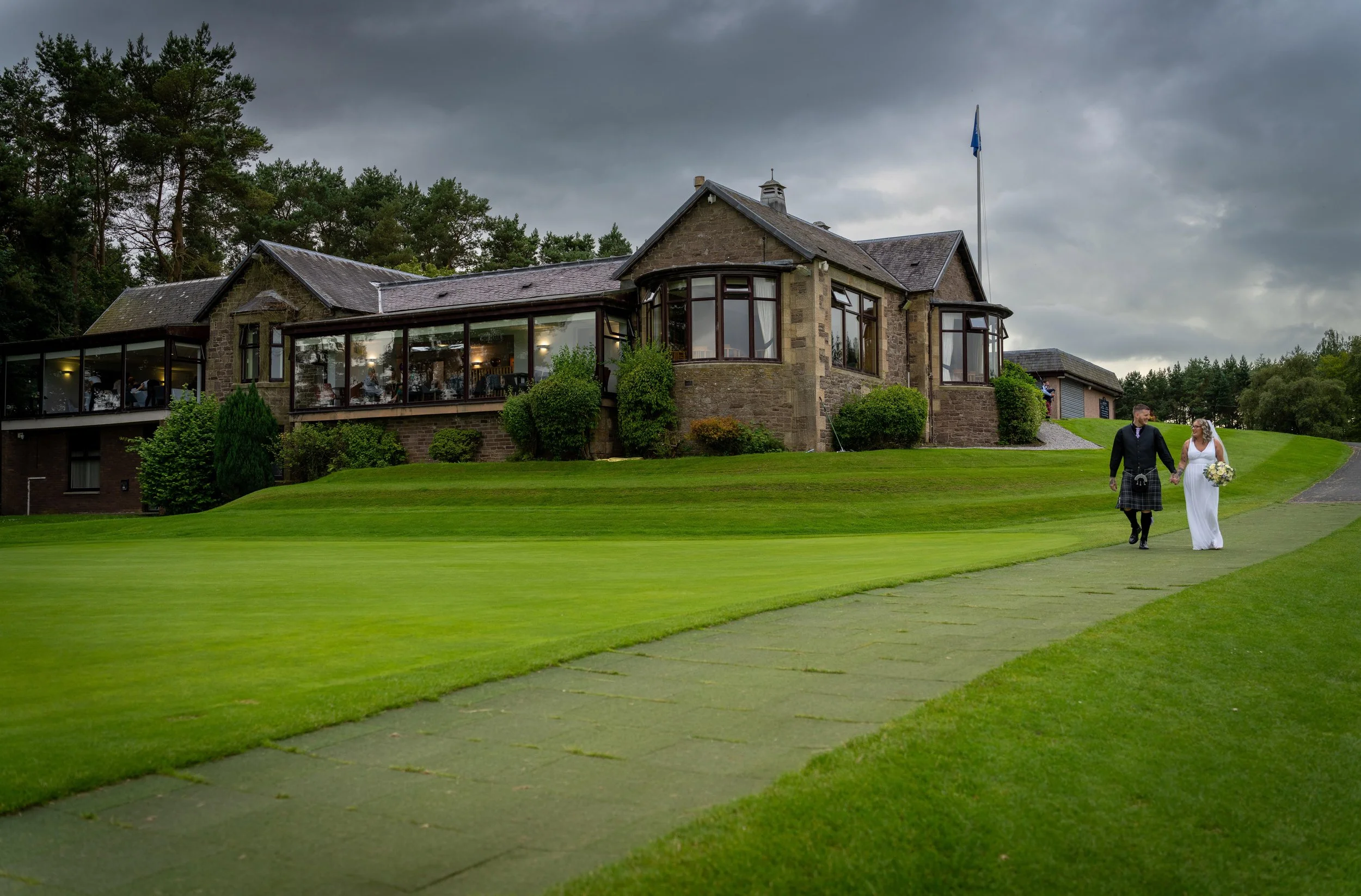 A bride and groom walking on a green golf course path in front of a large stone building with large windows, under a cloudy sky.