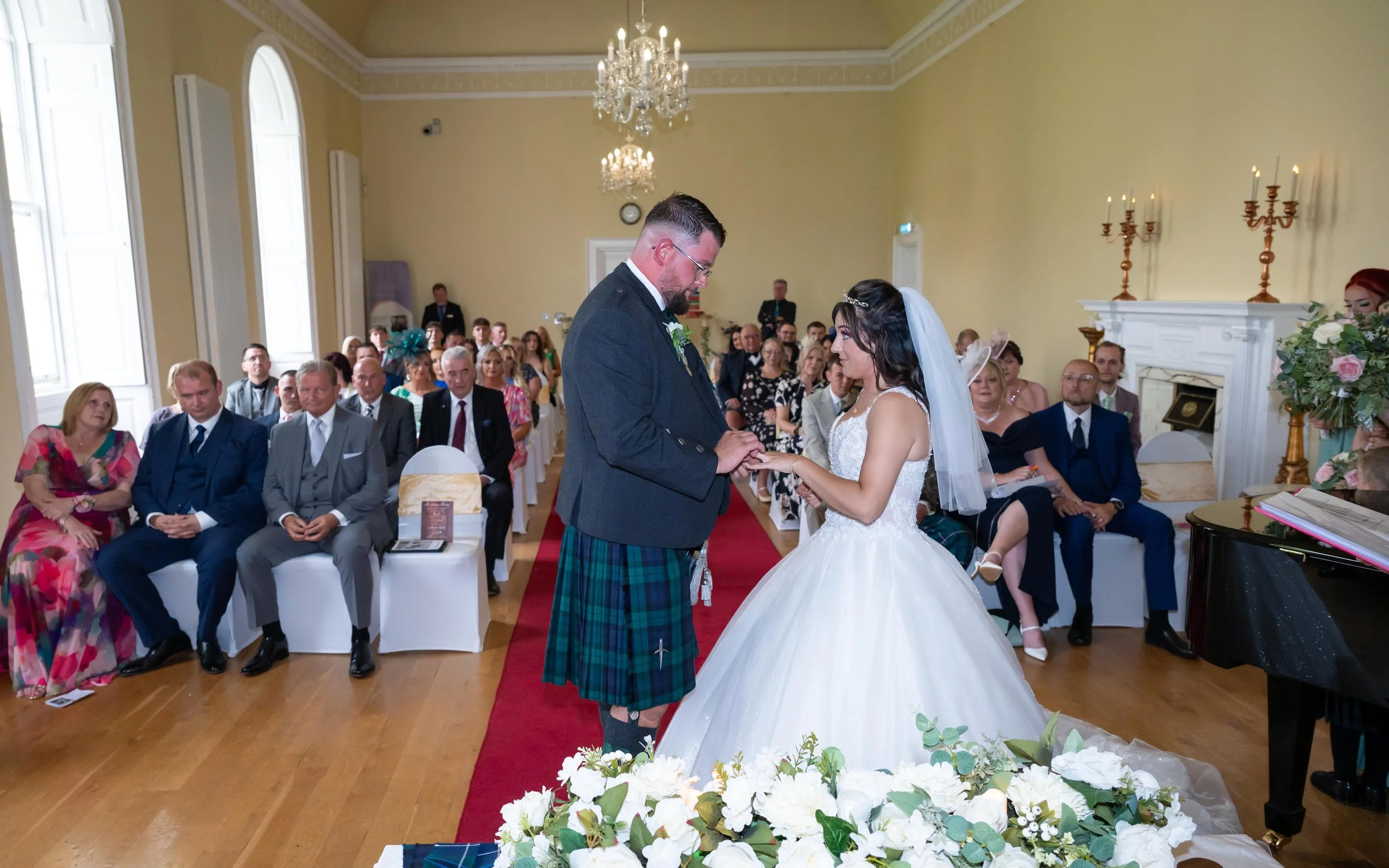 A wedding ceremony with a couple exchanging rings in a bright, elegant room. The bride is wearing a white gown and veil, and the groom is dressed in a kilt and jacket. Guests are seated around them, watching the bride and groom during the vows.