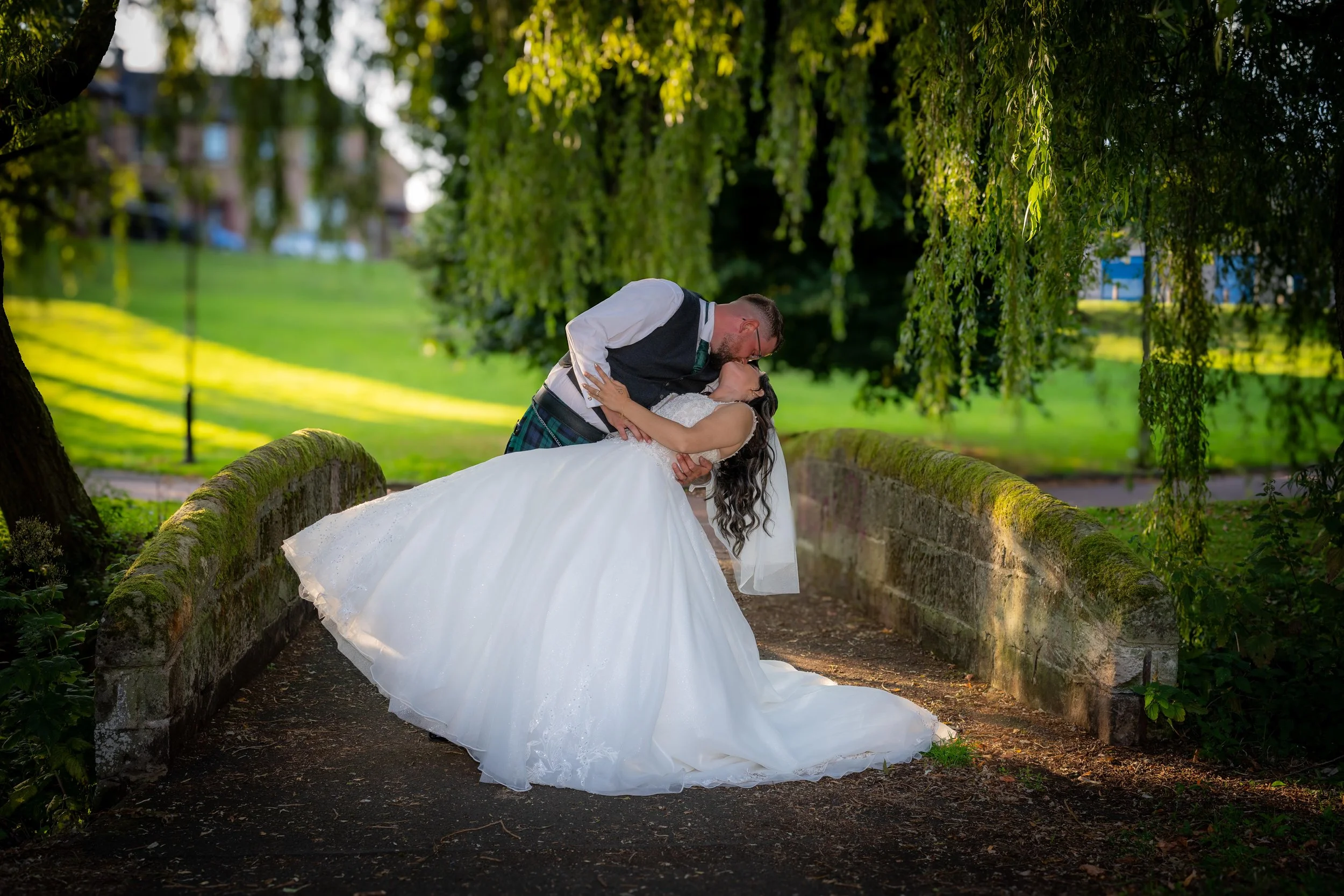 A bride and groom sharing a kiss on a small stone bridge in a park, with greenery and trees in the background.