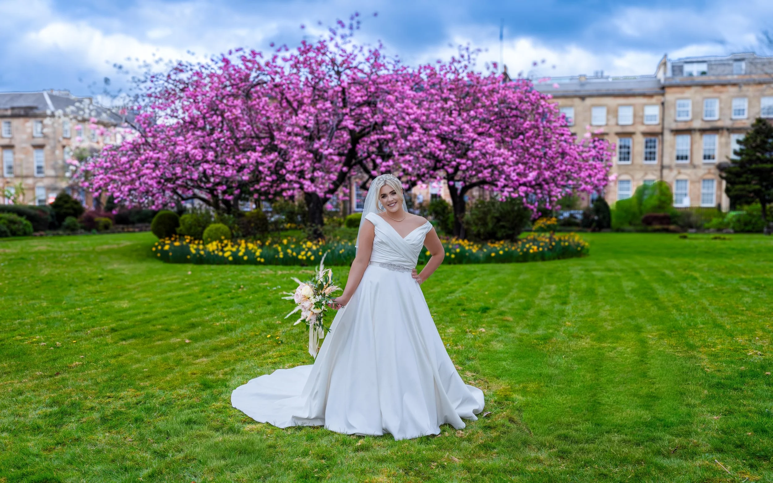 A bride in a white wedding gown holding a bouquet of flowers stands on a green lawn with a large pink flowering tree and buildings in the background.
