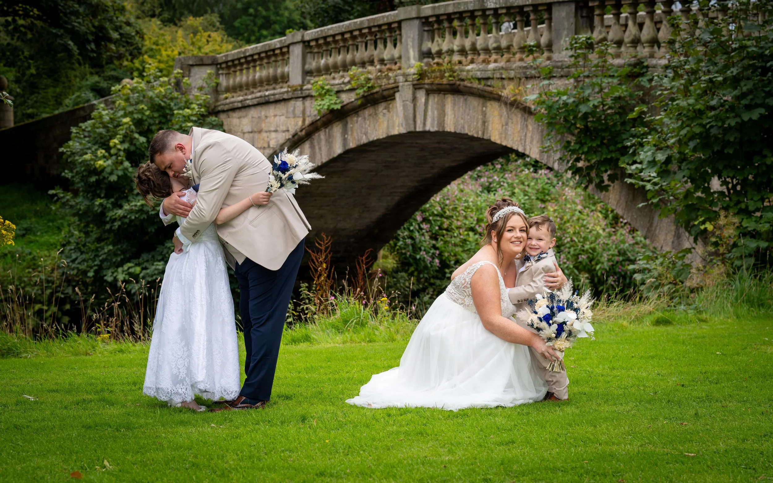 A wedding scene with two women in white wedding dresses, a man in a beige suit, and a young boy in a beige suit with a bow tie, holding bouquets and embracing on a grassy area near a stone bridge surrounded by greenery and flowers.