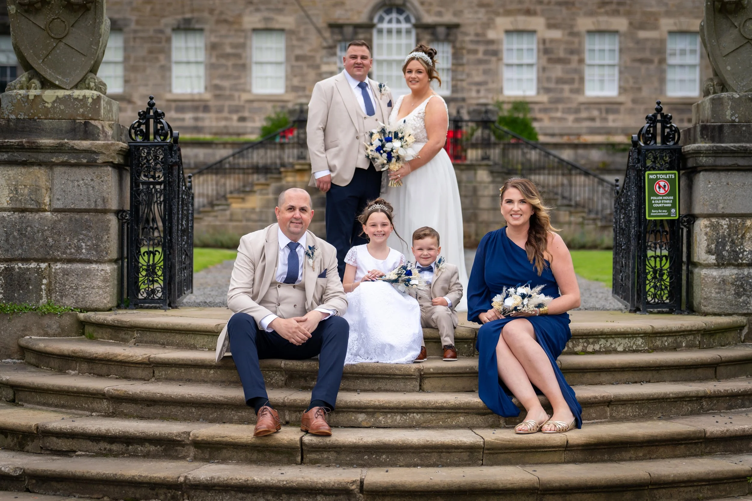 A wedding party poses on the steps of a historic building. The bride and groom stand at the top, while two children and two adults sit on the steps below.