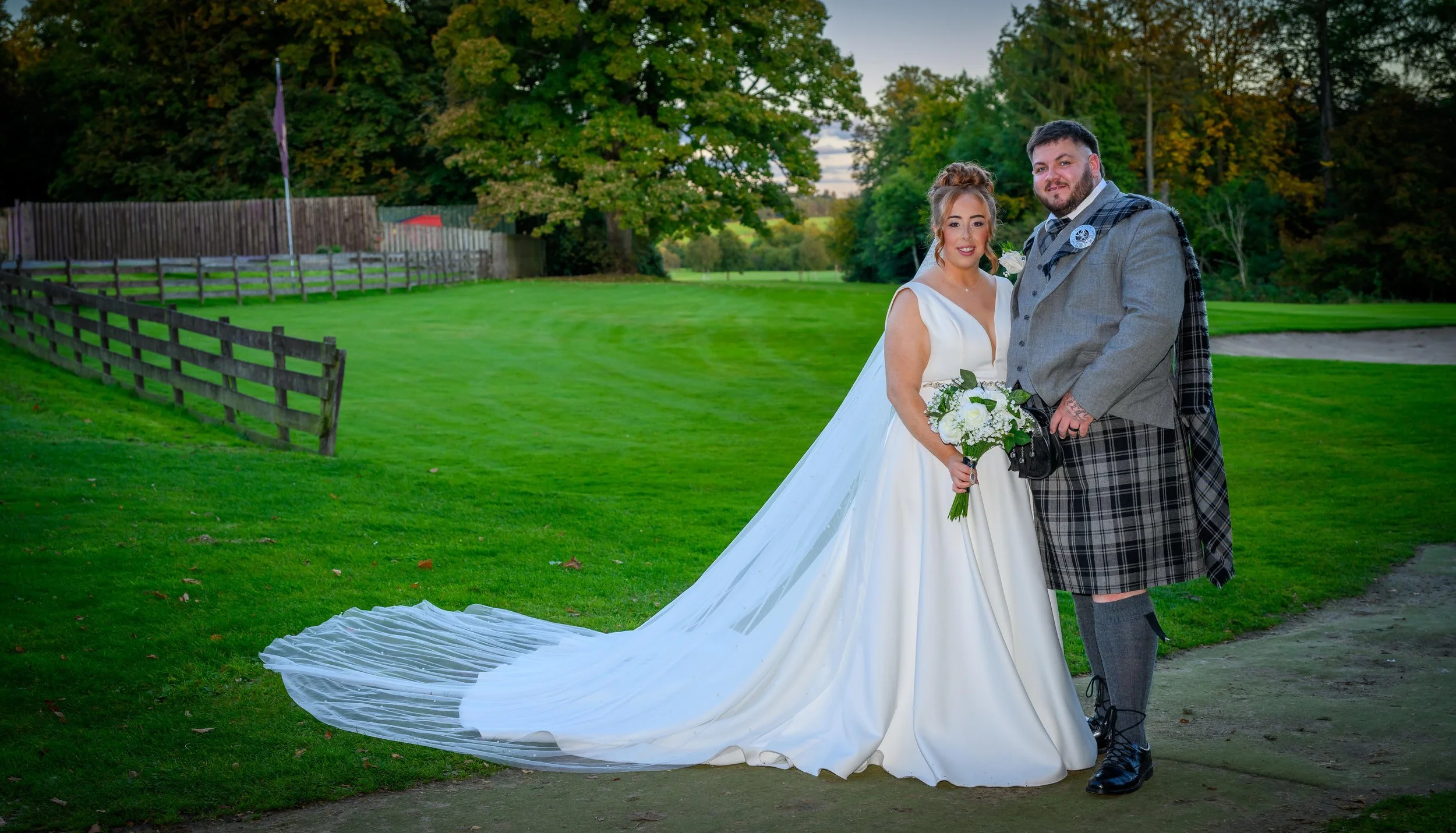 A bride and groom standing on a grassy area outdoors during daytime. The bride wears a white wedding gown and holds a bouquet of white flowers. The groom is dressed in traditional Scottish attire, including a kilt and a checked shawl. They are smilin