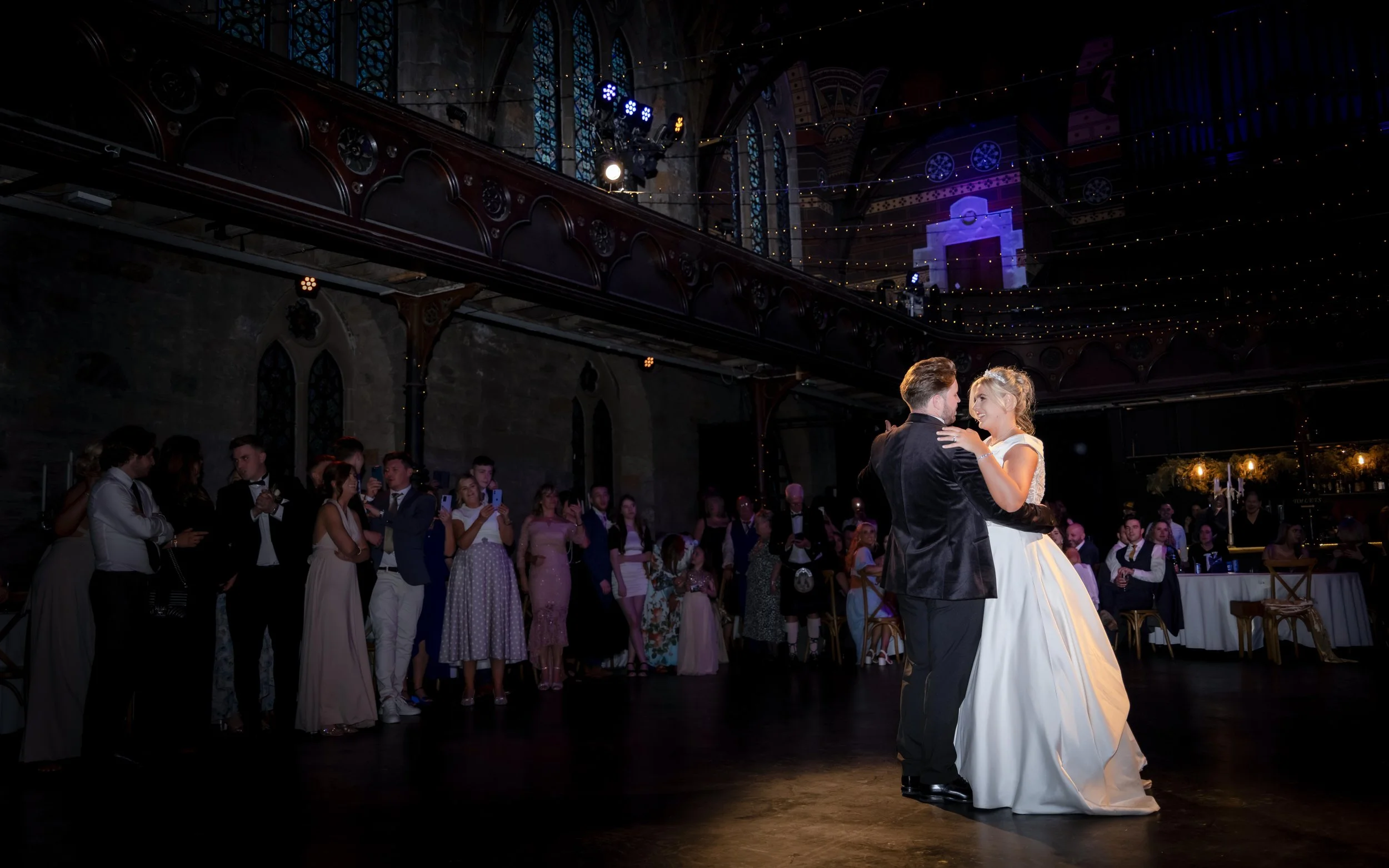 Bride and groom dancing during their wedding reception in a dimly lit, elegant hall with high ceiling, stained glass windows, and guests watching and taking photos.
