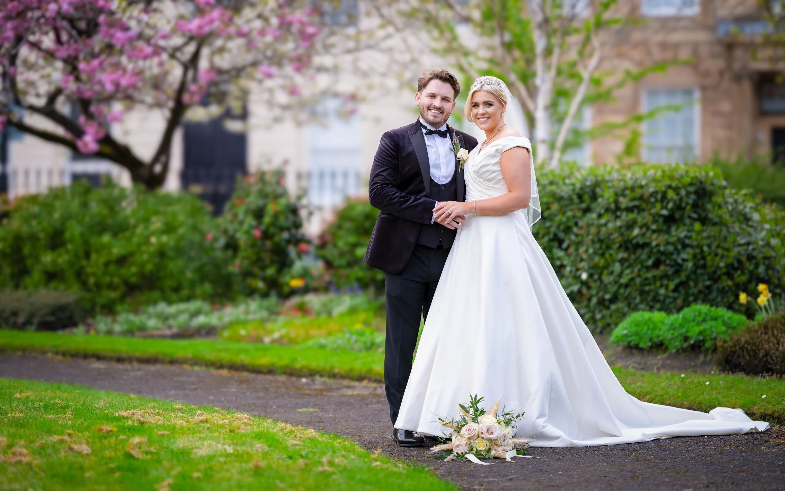 A newlywed couple in wedding attire standing on a garden path, smiling and holding hands, with a wedding bouquet on the ground in front of them, surrounded by blooming trees and greenery.