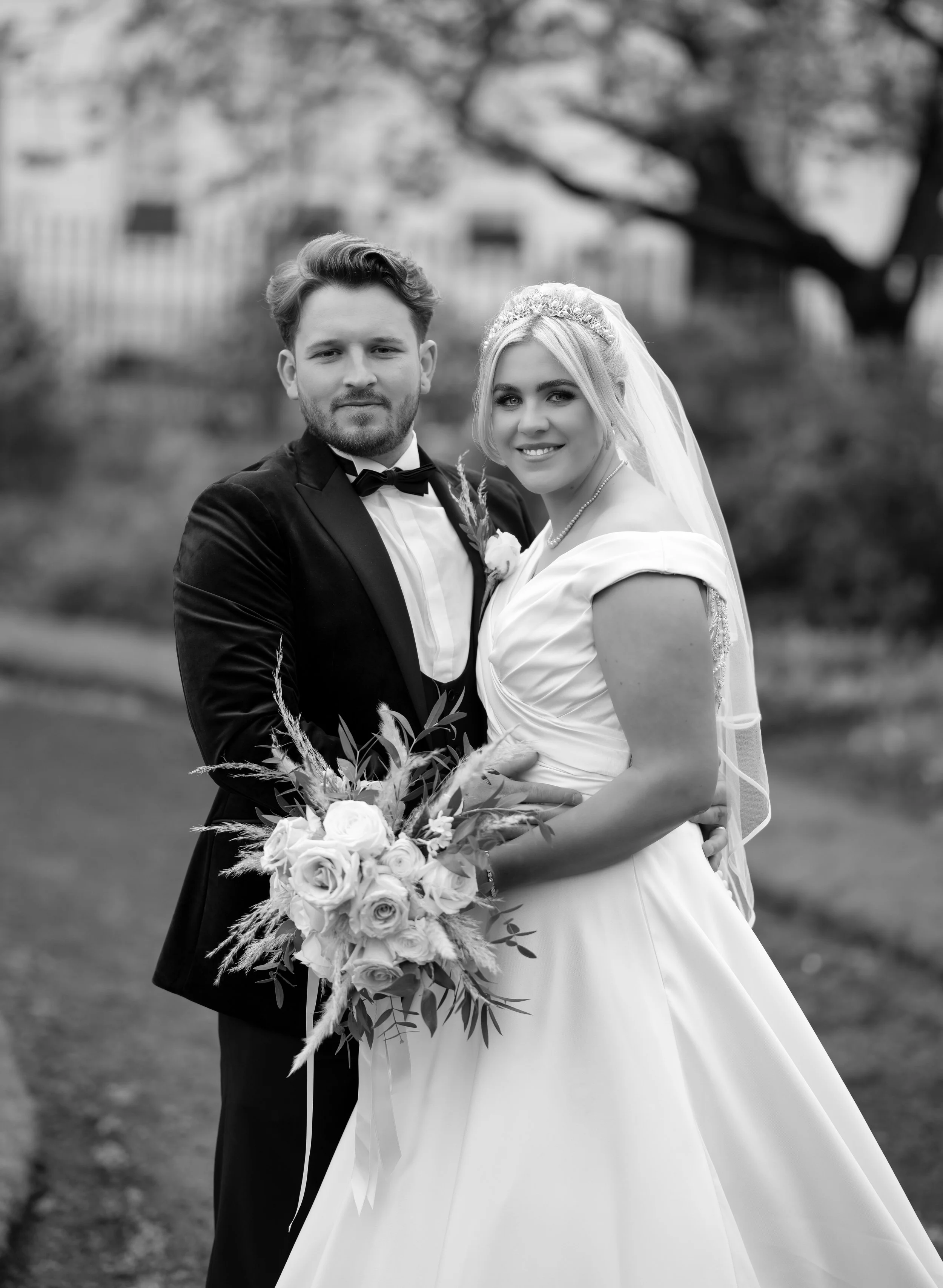 Black and white photo of a bride and groom on their wedding day outdoors. The bride is smiling, holding a bouquet of roses, and wearing a wedding gown with a veil. The groom is wearing a tuxedo with a bow tie. They are standing close together in fron