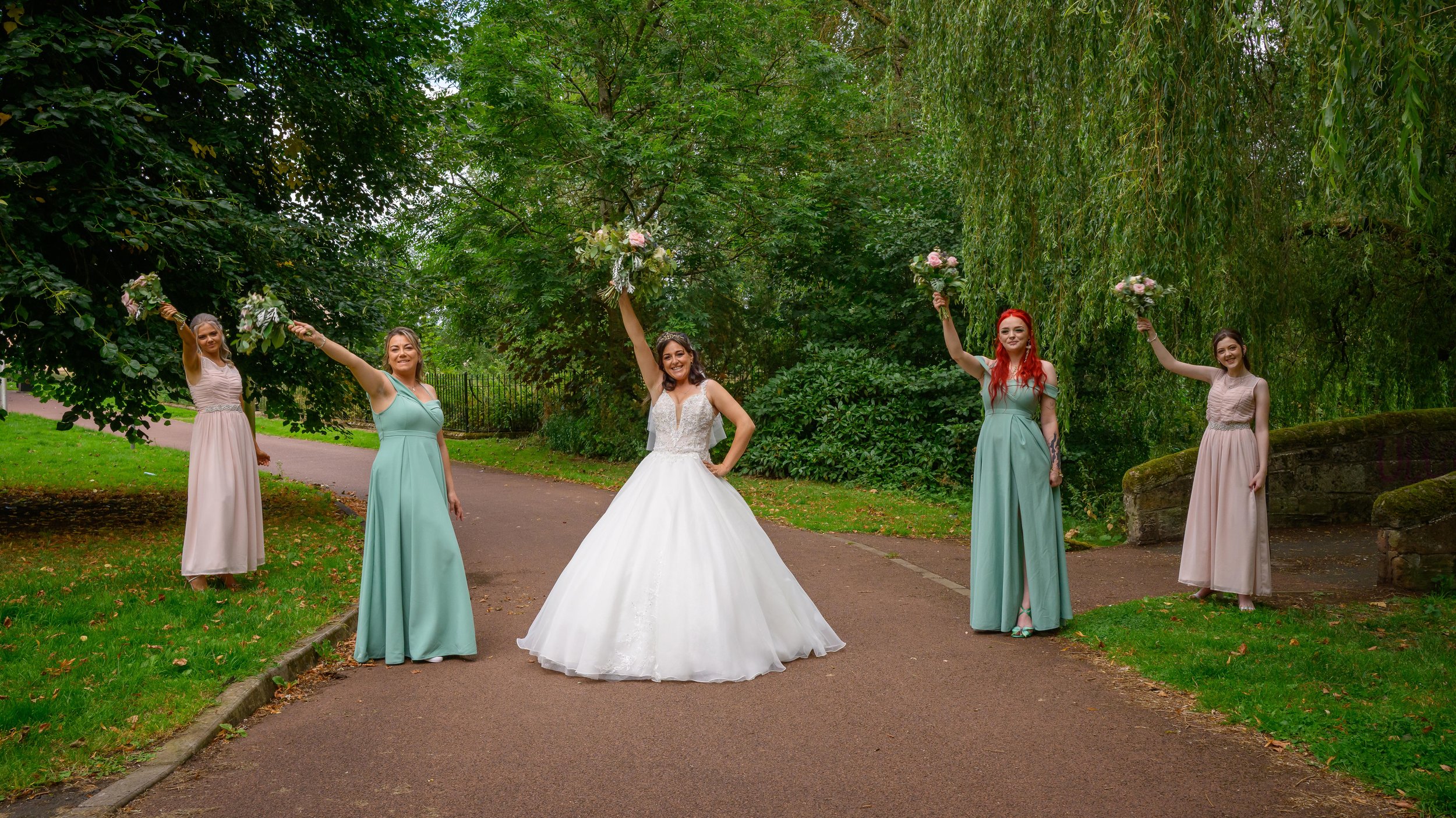 A bride with dark hair in a white wedding gown, holding a bouquet, standing on a path in a park with four bridesmaids in pastel dresses, all holding bouquets and smiling.