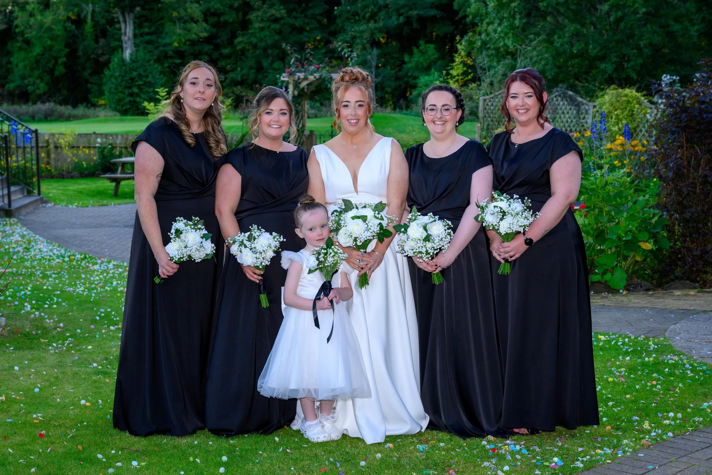 A group of six women and one young girl standing outdoors on a grassy area with trees and bushes in the background. They are dressed for a wedding, with the bride in a white gown and the others in black dresses. They are holding bouquets of white flo