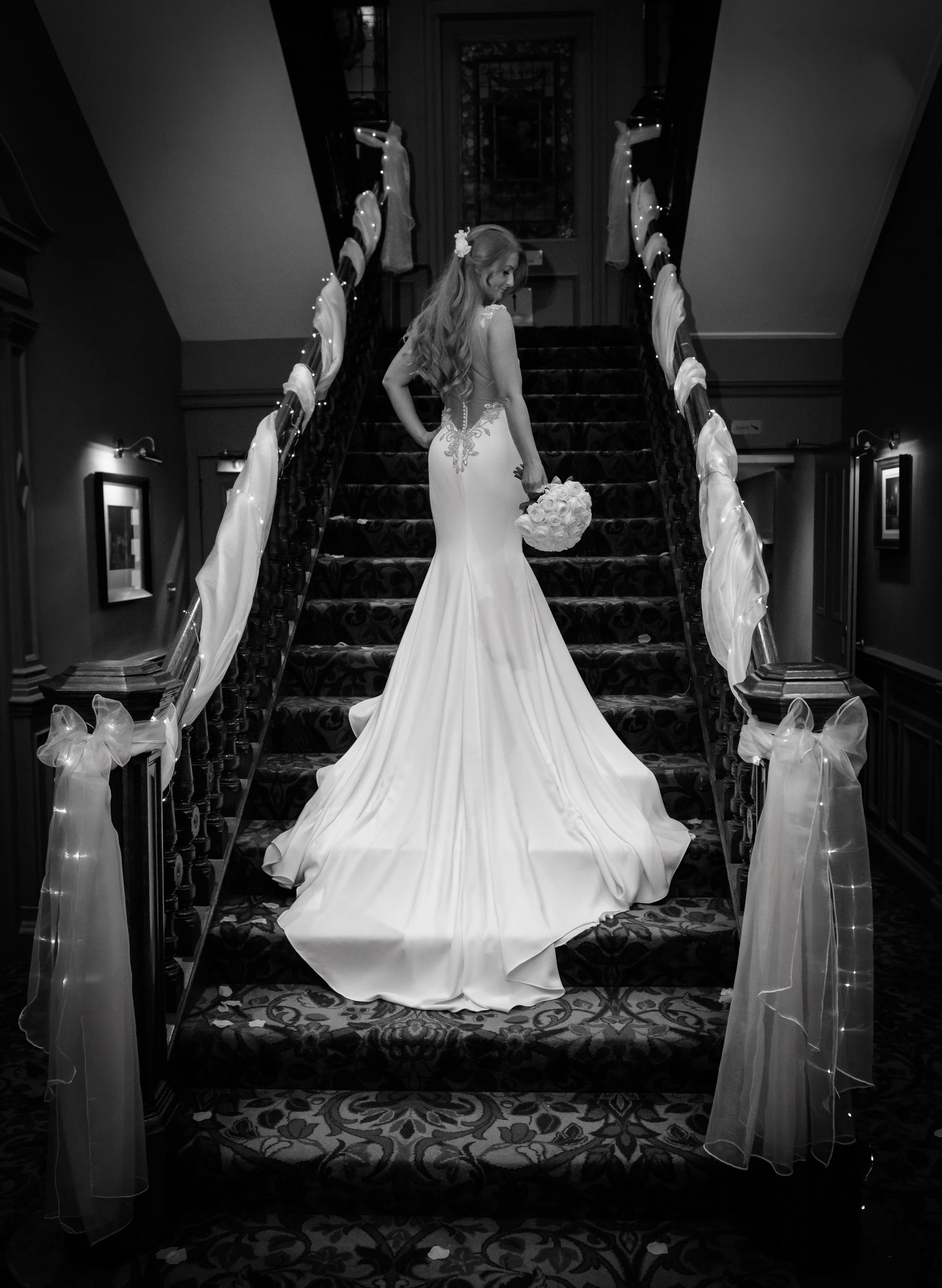 A bride in a long, elegant wedding dress holding a bouquet of roses on a decorated staircase inside a building.