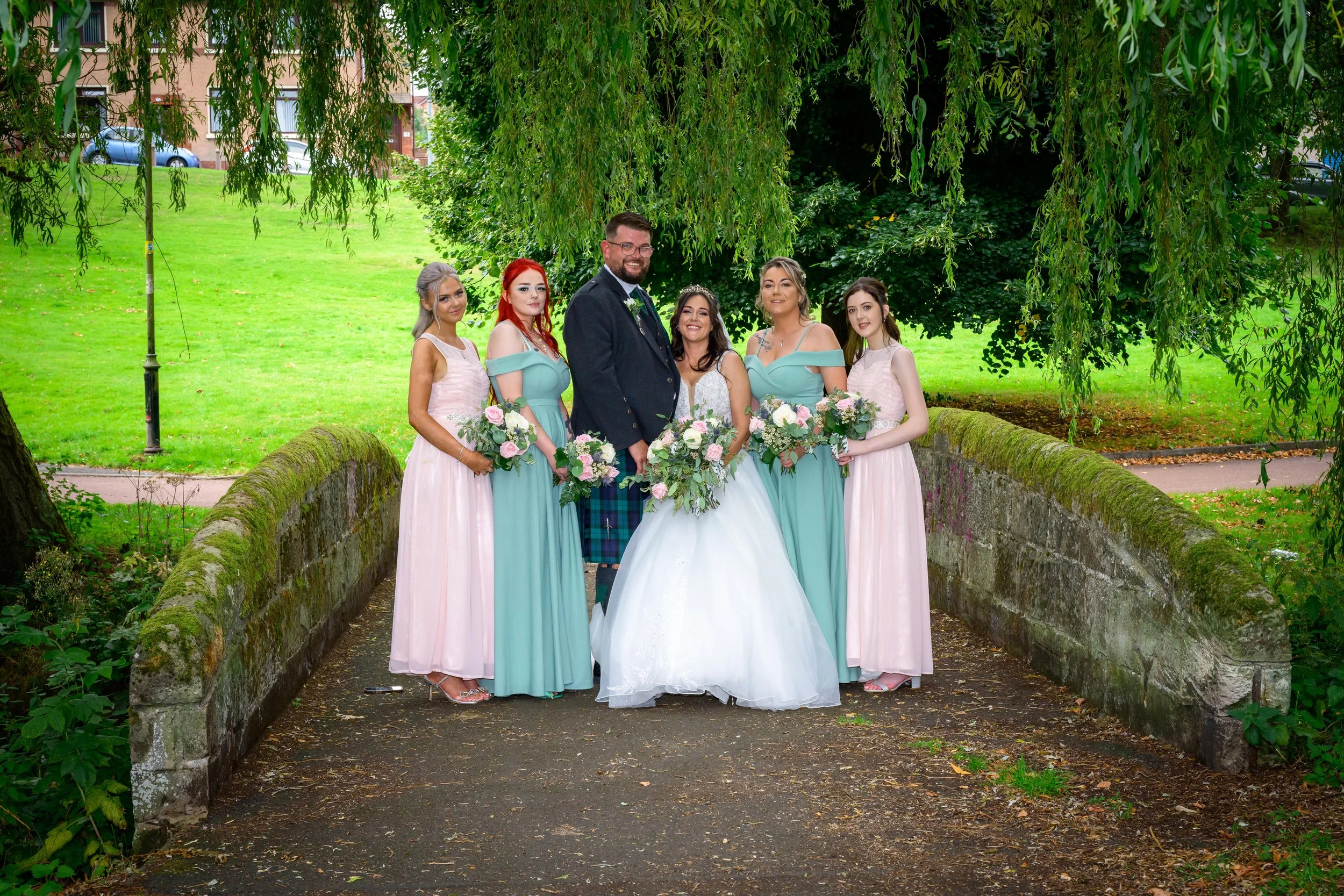 A wedding group photo outdoors on a stone bridge, featuring a bride in a white gown, a groom in a dark jacket and kilt, and four bridesmaids in pastel dresses, all holding bouquets of flowers, surrounded by lush greenery.
