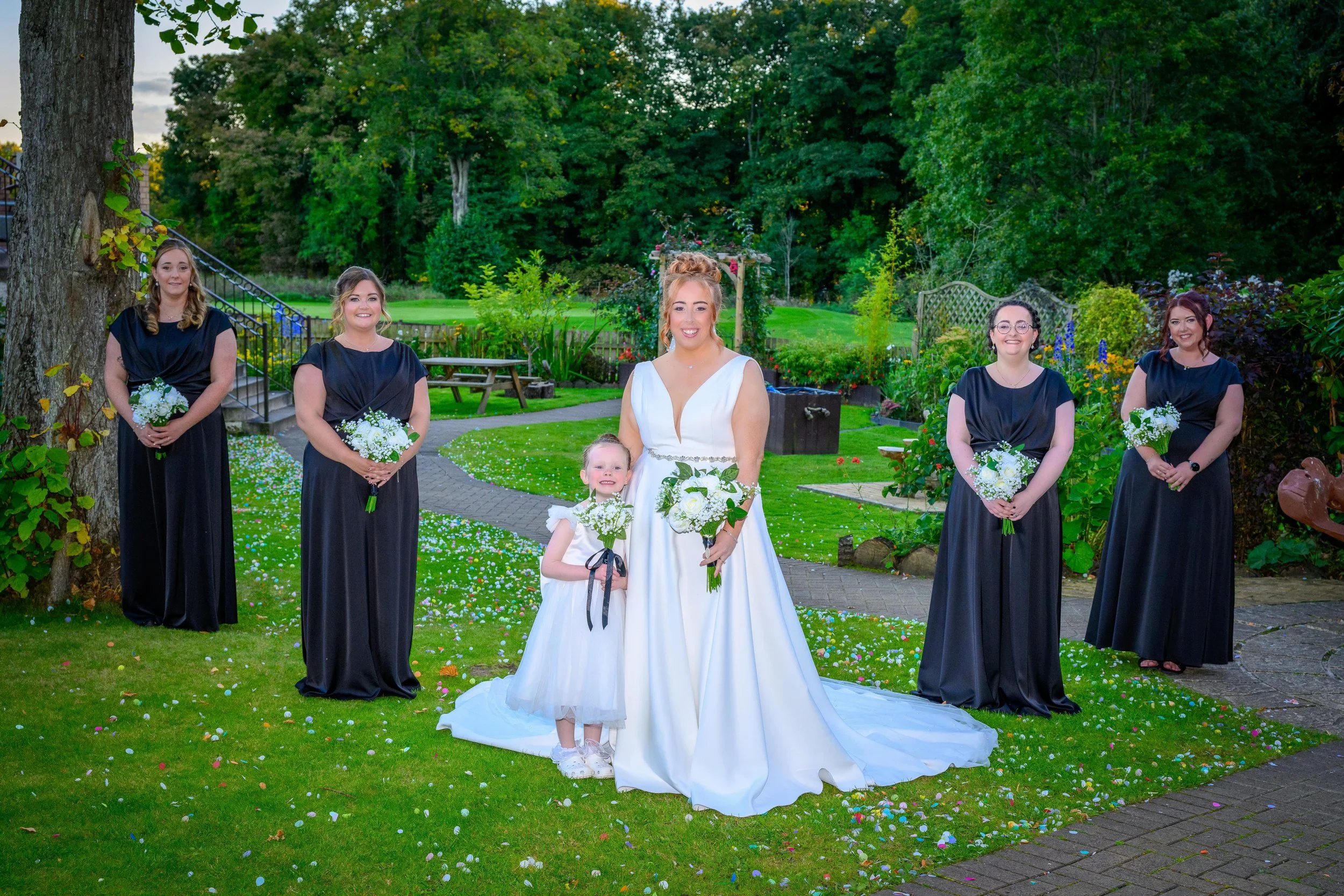 A bride in a white wedding gown with a young girl in a white dress, surrounded by five bridesmaids dressed in black gowns, at an outdoor garden wedding ceremony. The bride and girl hold bouquets of white flowers, and the setting includes greenery, tr