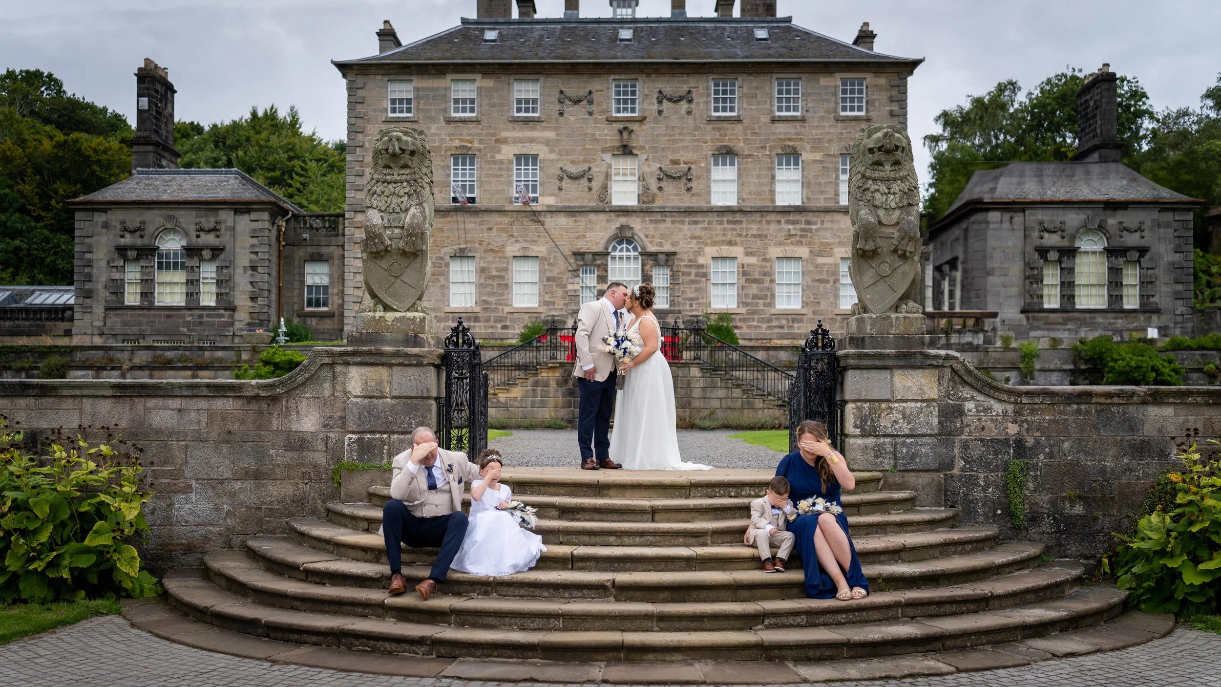 A wedding couple kisses on the steps of a historic stone building with statues of lions on pillars and a group of family and friends sitting on the steps, covering their faces, celebrating the wedding outside.
