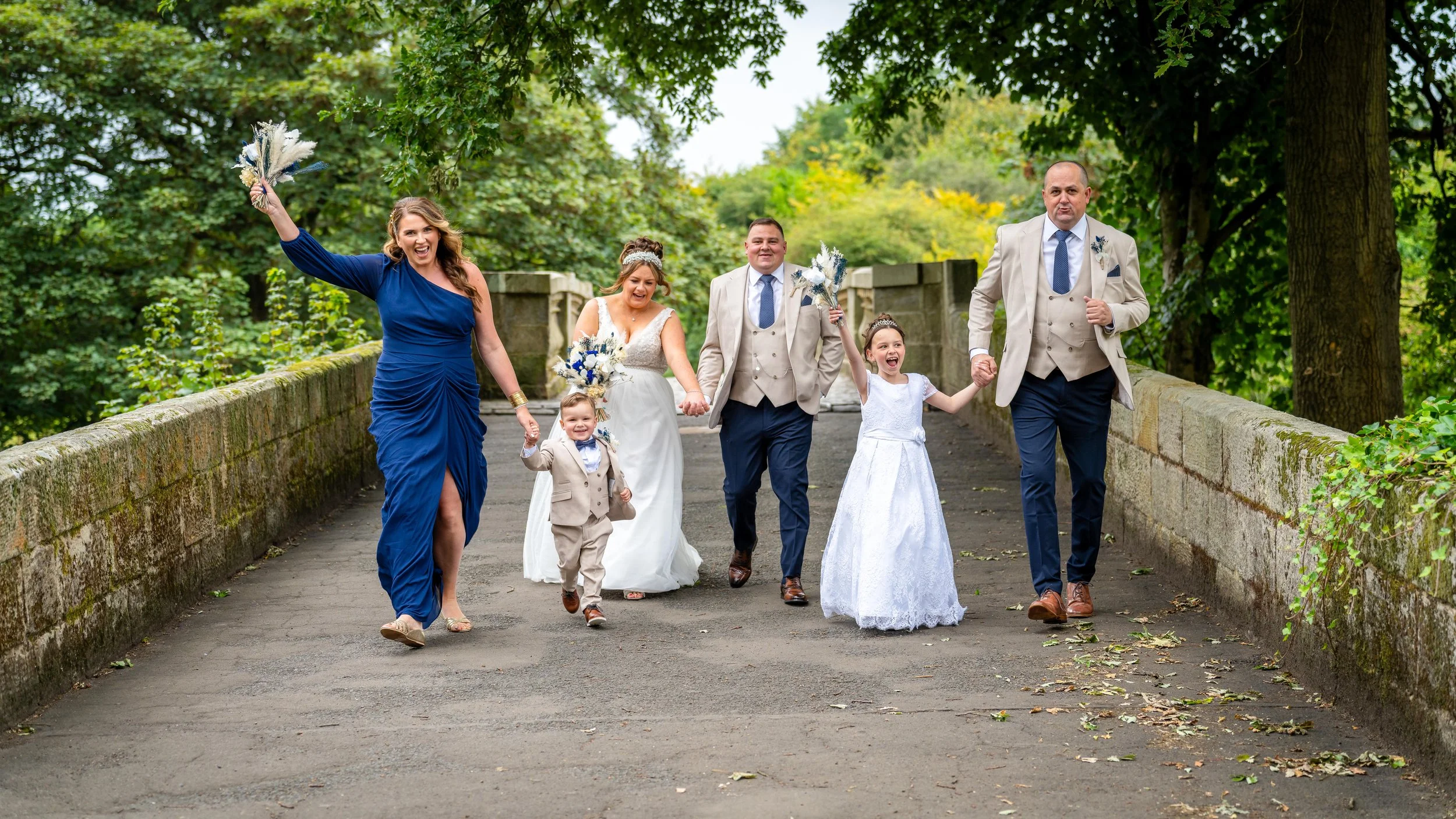 A wedding party of six walking on a bridge surrounded by trees. The bride is wearing a white wedding dress, the groom and a man are in beige suits, a woman is in a long blue dress, and two children are in white dresses and beige suits. They are smili