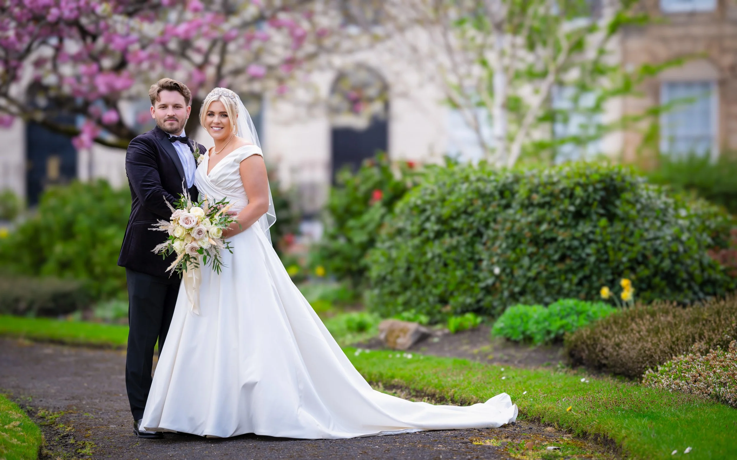 Bride and groom in wedding attire standing on a garden pathway with blooming trees and shrubs in the background.