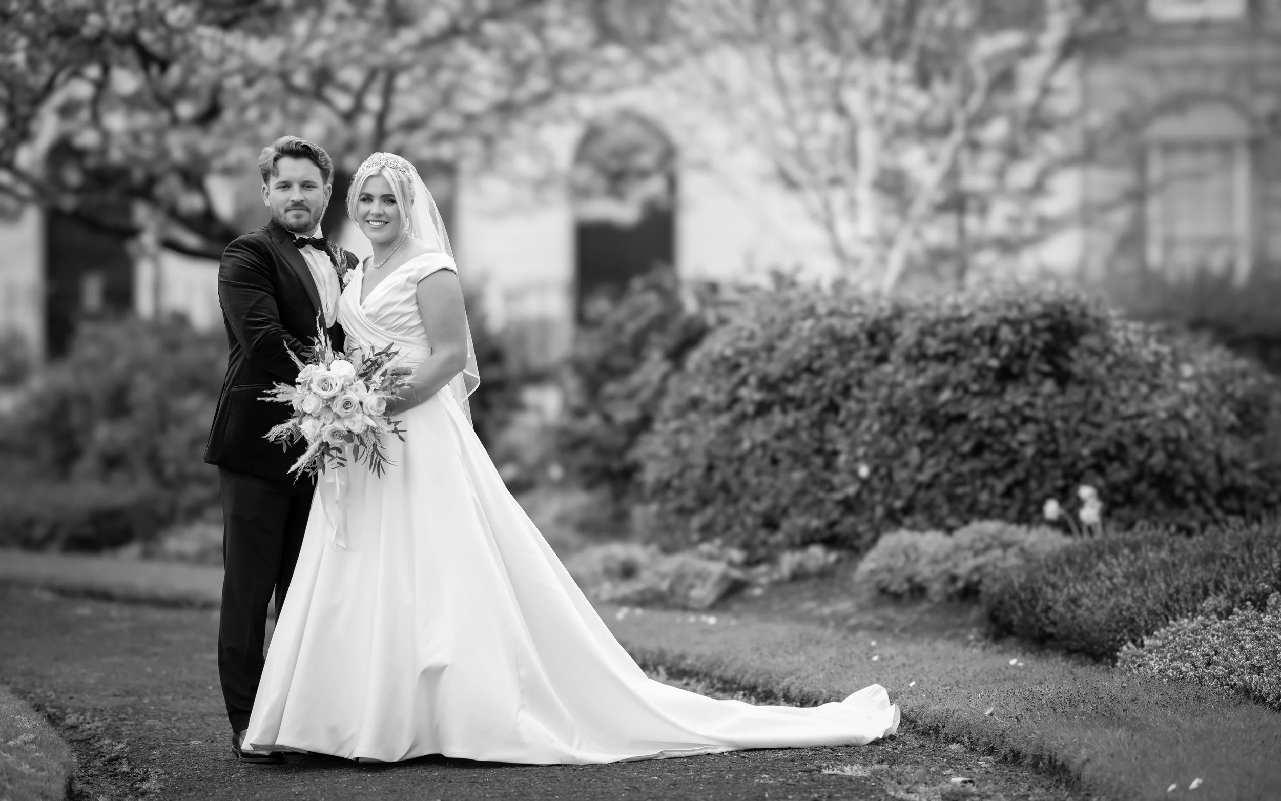 A black and white photograph of a bride and groom standing together outdoors in a garden, with wedding attire. The bride is holding a bouquet and smiling, while the groom is standing beside her with a serious expression. Trees and bushes are visible 