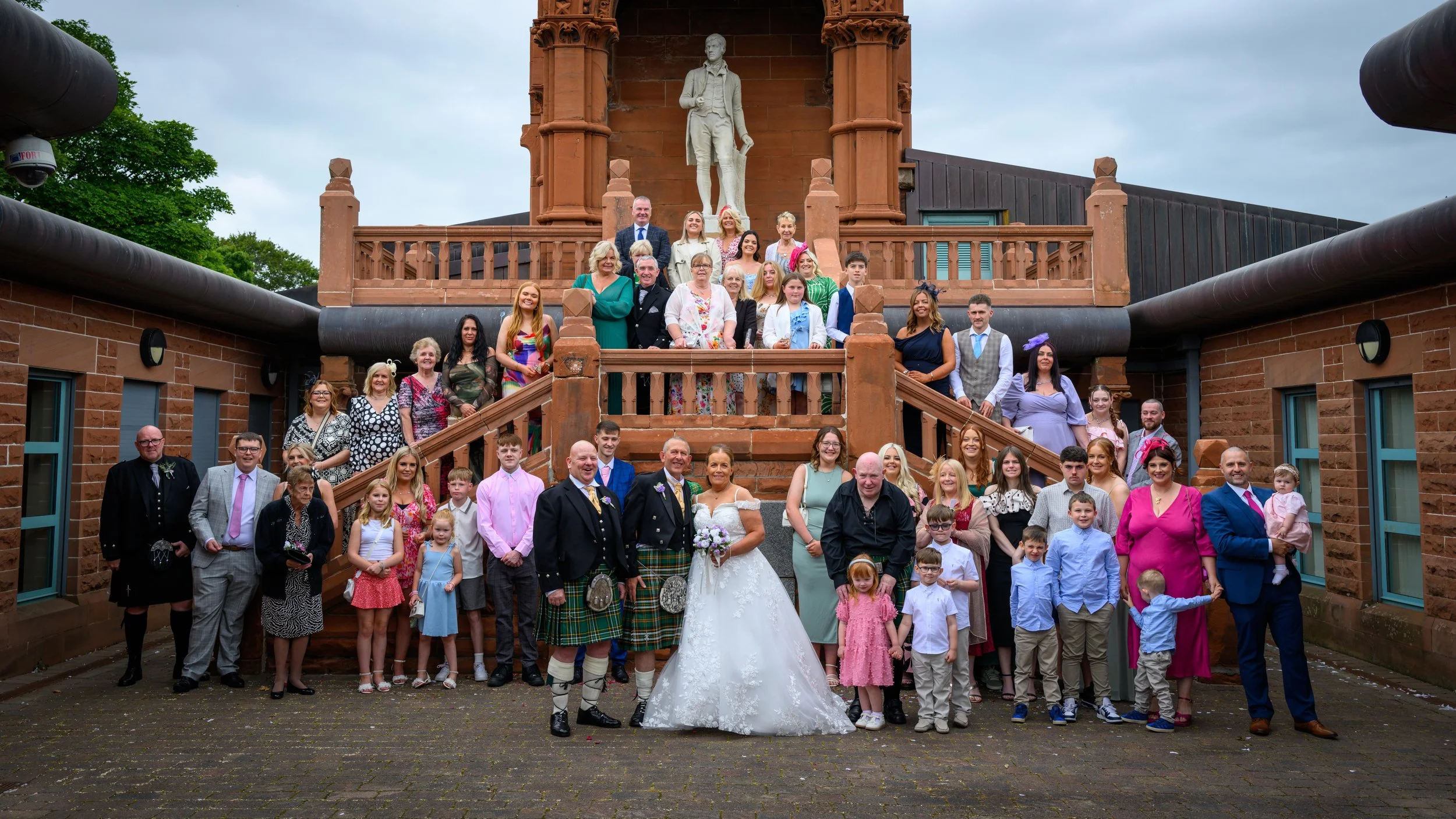 A large group of people, including a bride and groom, gathers for a wedding photo outside a historic red sandstone building with a statue of a man above the staircase. The scene is overcast.