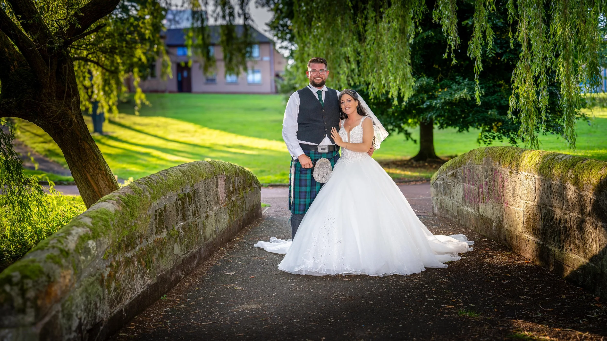 Bride and groom dressed in wedding attire standing on a small stone bridge in a park with lush green trees and grass.