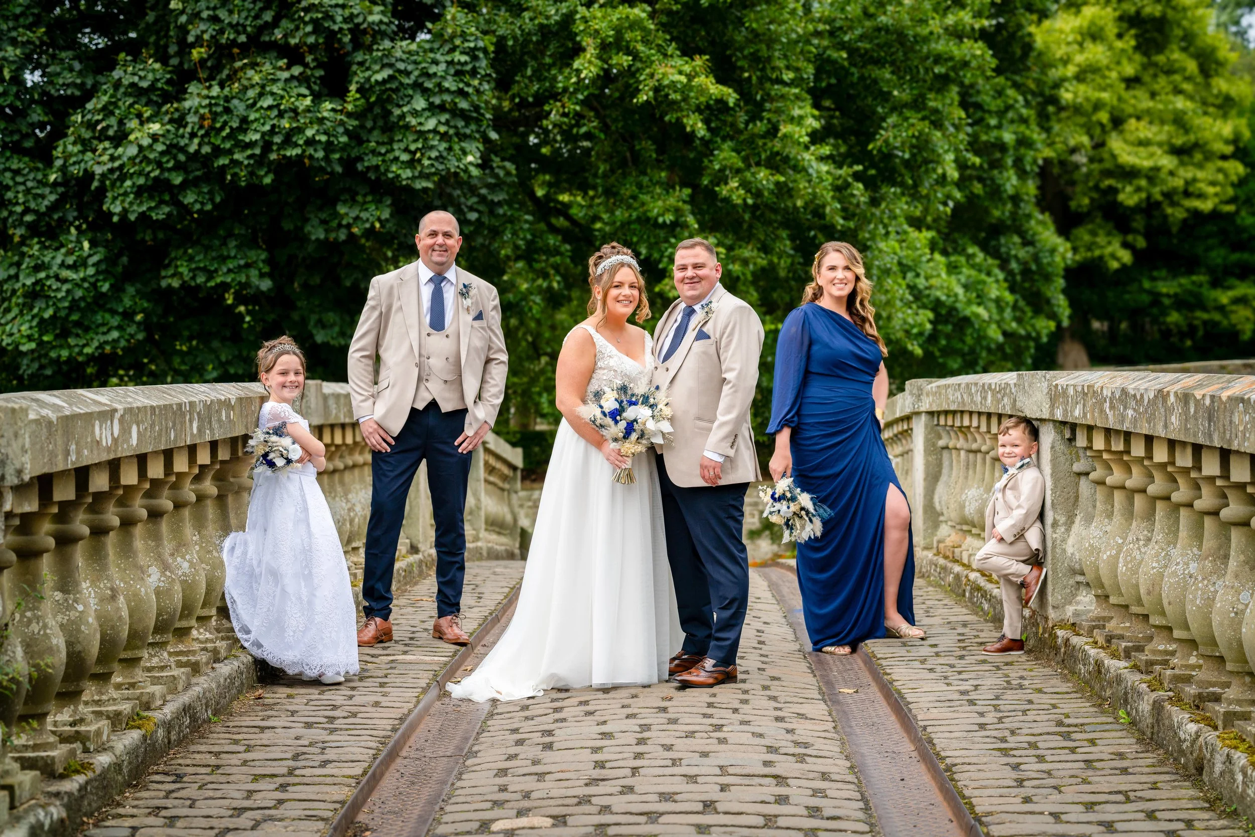 A wedding party with six people standing on a cobblestone bridge, surrounded by green trees. The bride and groom are in the center, smiling and holding bouquets. Two children and two adults are on the sides, also smiling; the children are dressed in 