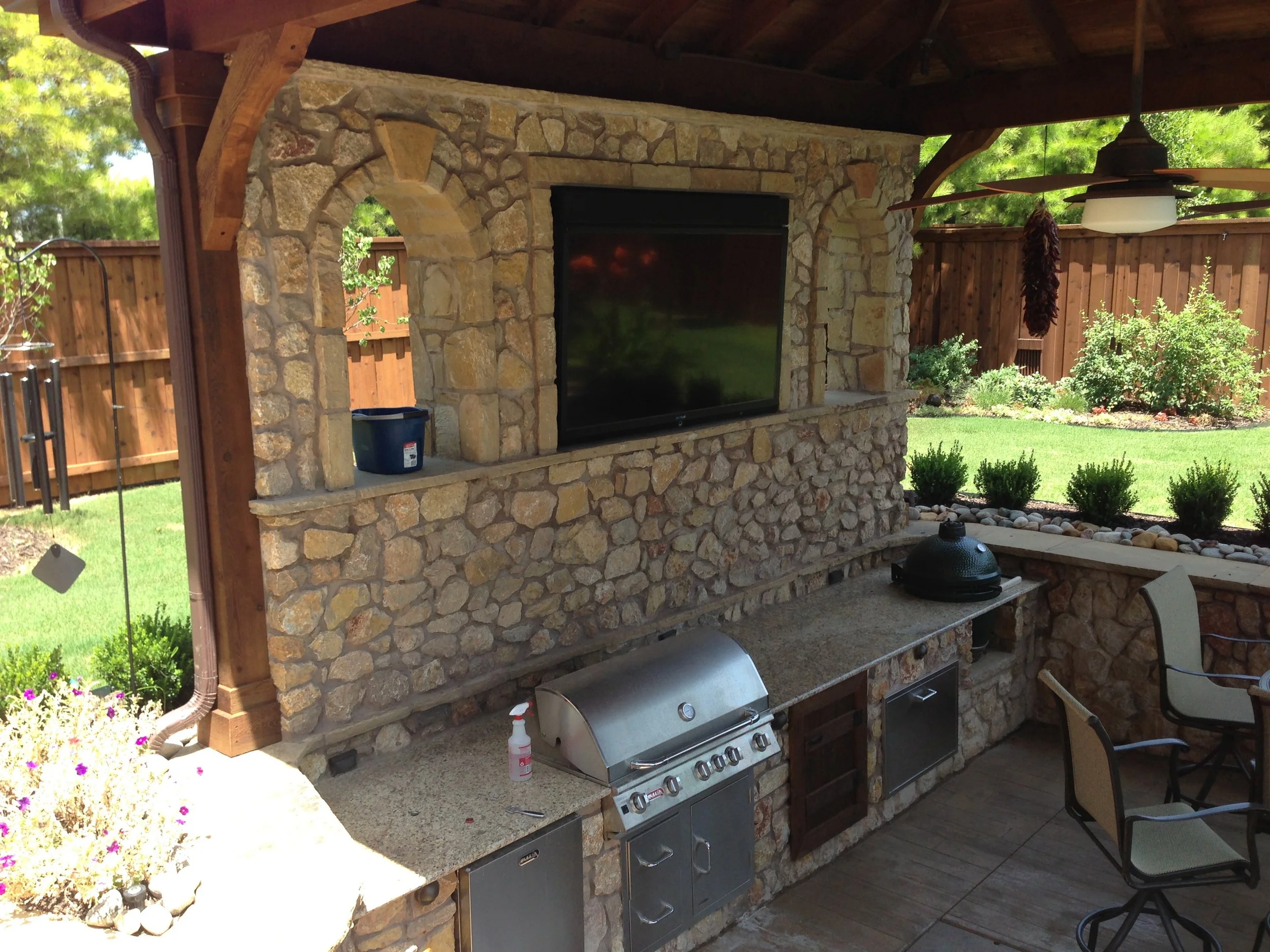 An outdoor kitchen with a stone countertop, stainless steel grill, black ceramic grill, and a TV mounted on a stone wall, under a wooden roof. There are chairs, a ceiling fan, and a landscaped backyard with plants and a wooden fence.