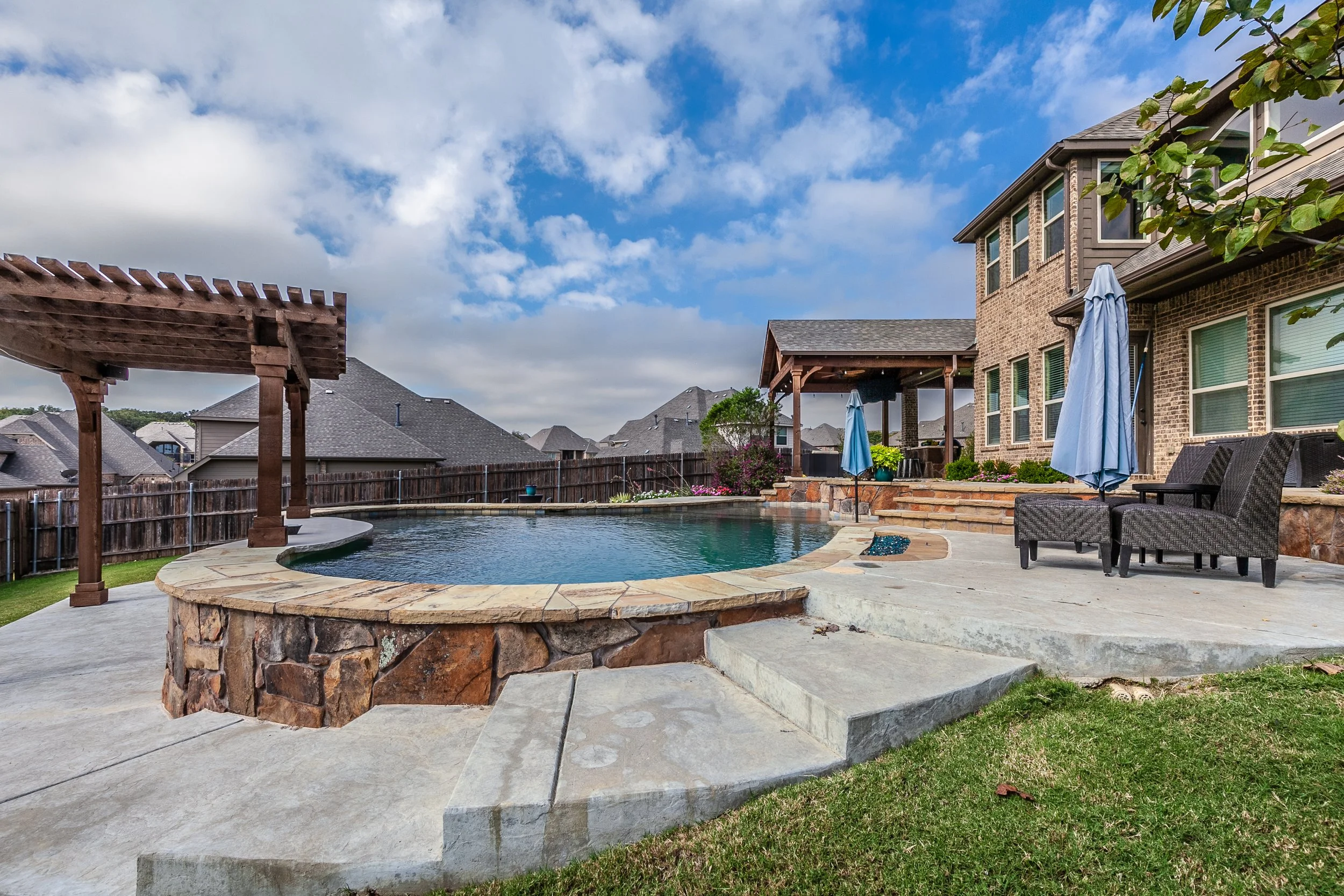 Backyard with an in-ground pool, stone border, poolside furniture, umbrellas, and a covered patio under a blue sky with clouds.
