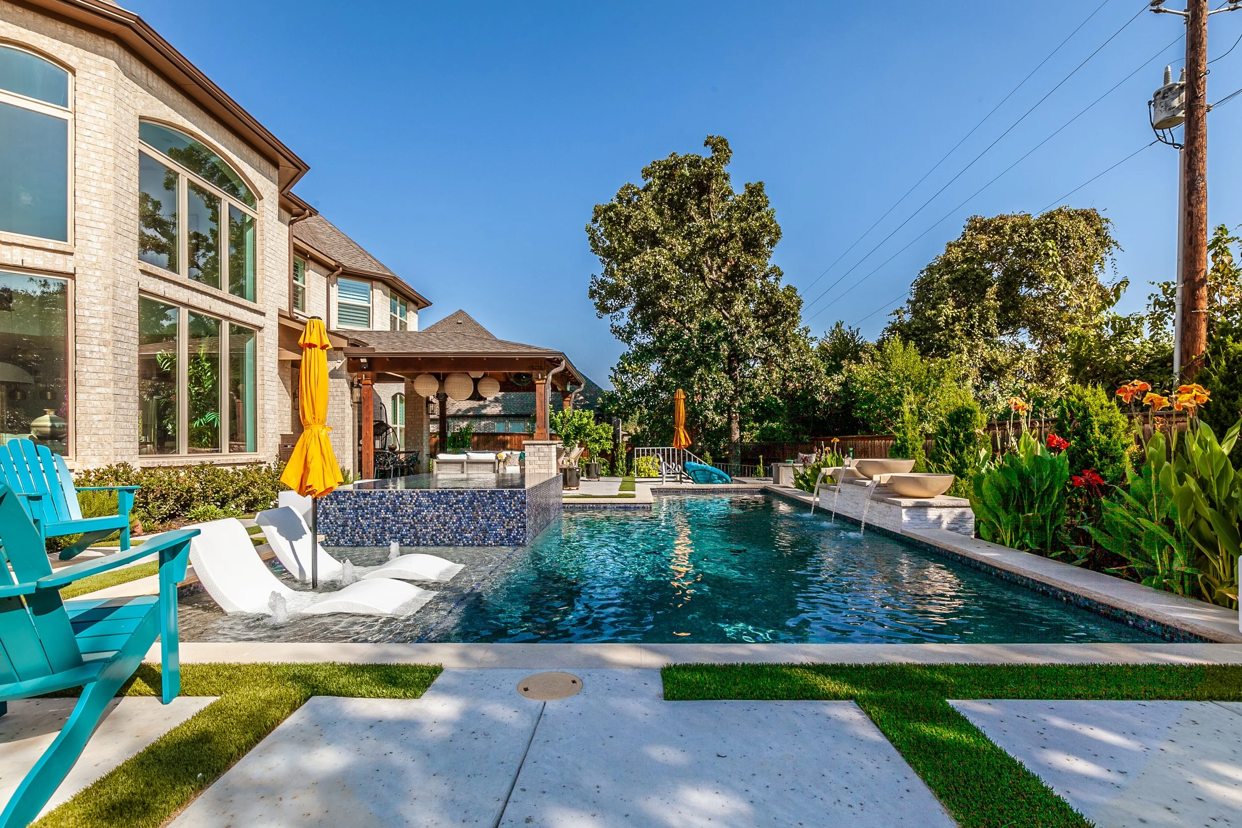 A backyard with a swimming pool, surrounded by patio furniture, umbrellas, trees, and plants, with a house in the background under a clear blue sky.