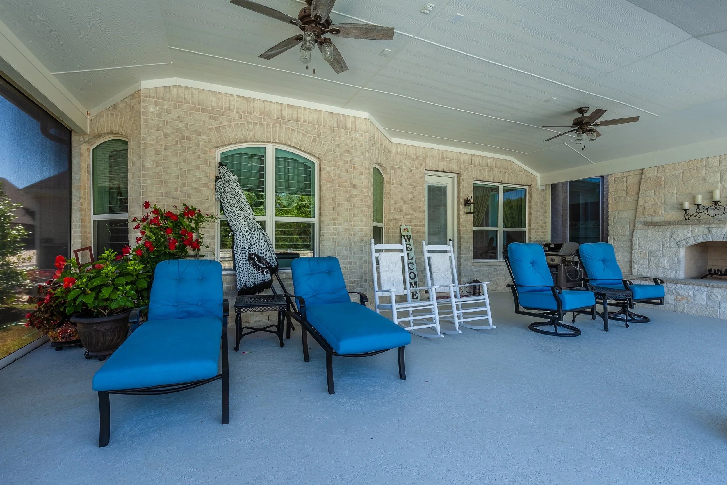 Covered back porch with blue cushioned chairs, white rocking chairs with a welcoming sign, a grill, and decorative plants in pots, enclosed by brick and stone walls, with fans and outdoor fireplace.