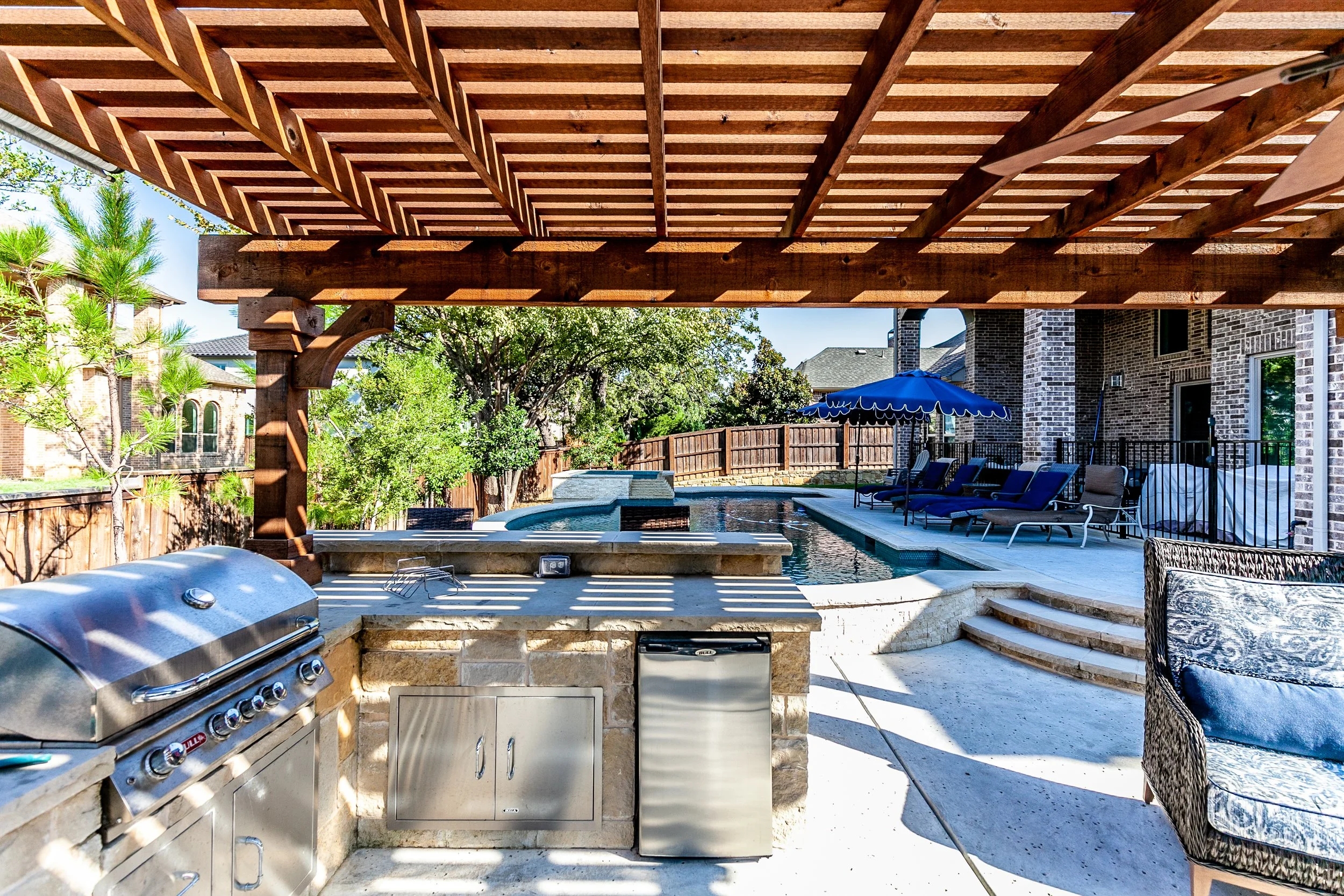 Outdoor backyard pool area with a covered patio, built-in grill, and lounge chairs under a blue umbrella.