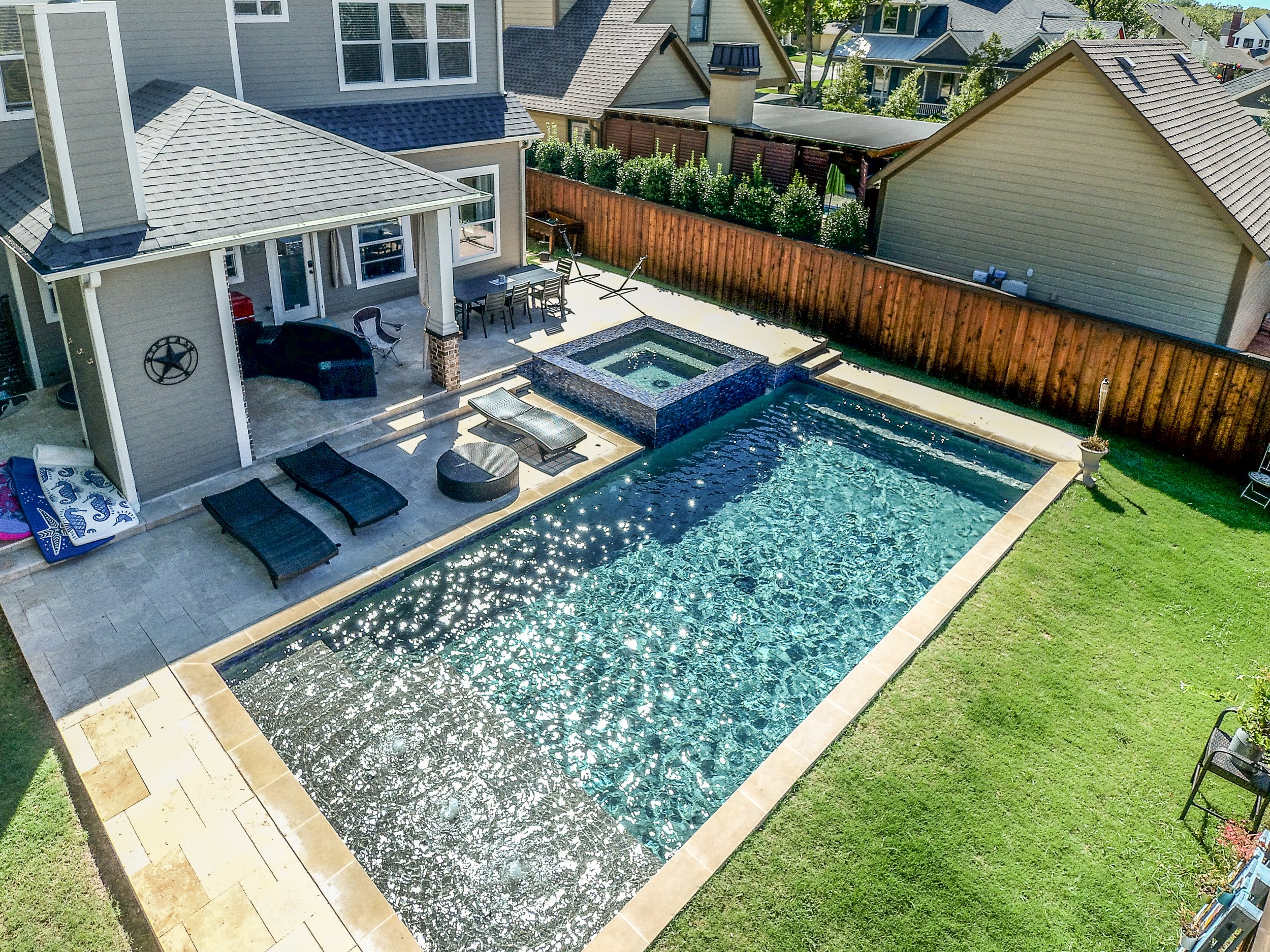 A backyard with a swimming pool, hot tub, patio furniture, and adjacent house with a deck.