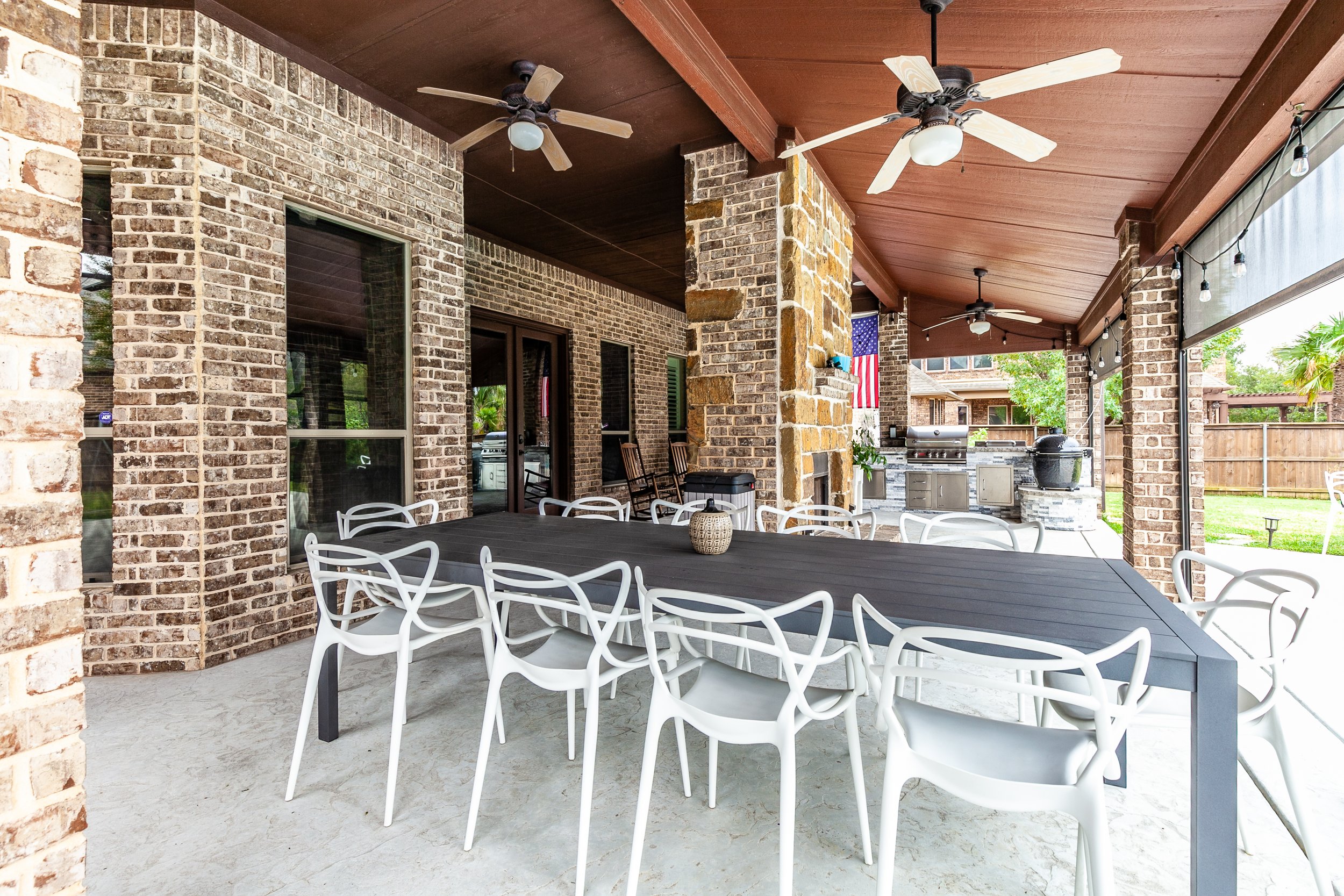 Covered outdoor patio with a large black dining table surrounded by white chairs, ceiling fans, an outdoor grill, a stone fireplace, an American flag, and a view of a fenced backyard with grass and trees.