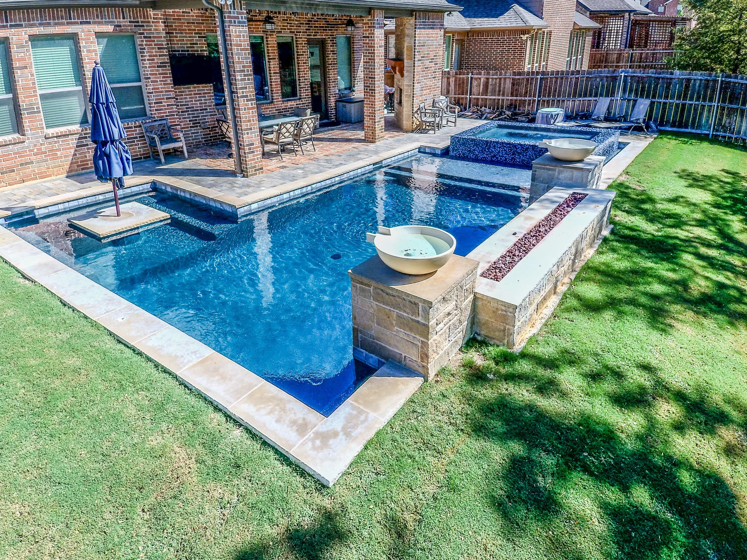 Backyard with a swimming pool, patio furniture, and a house with brick exterior. The pool has a small waterfall feature, and two large planters are placed on brick pillars at opposite ends of the pool.
