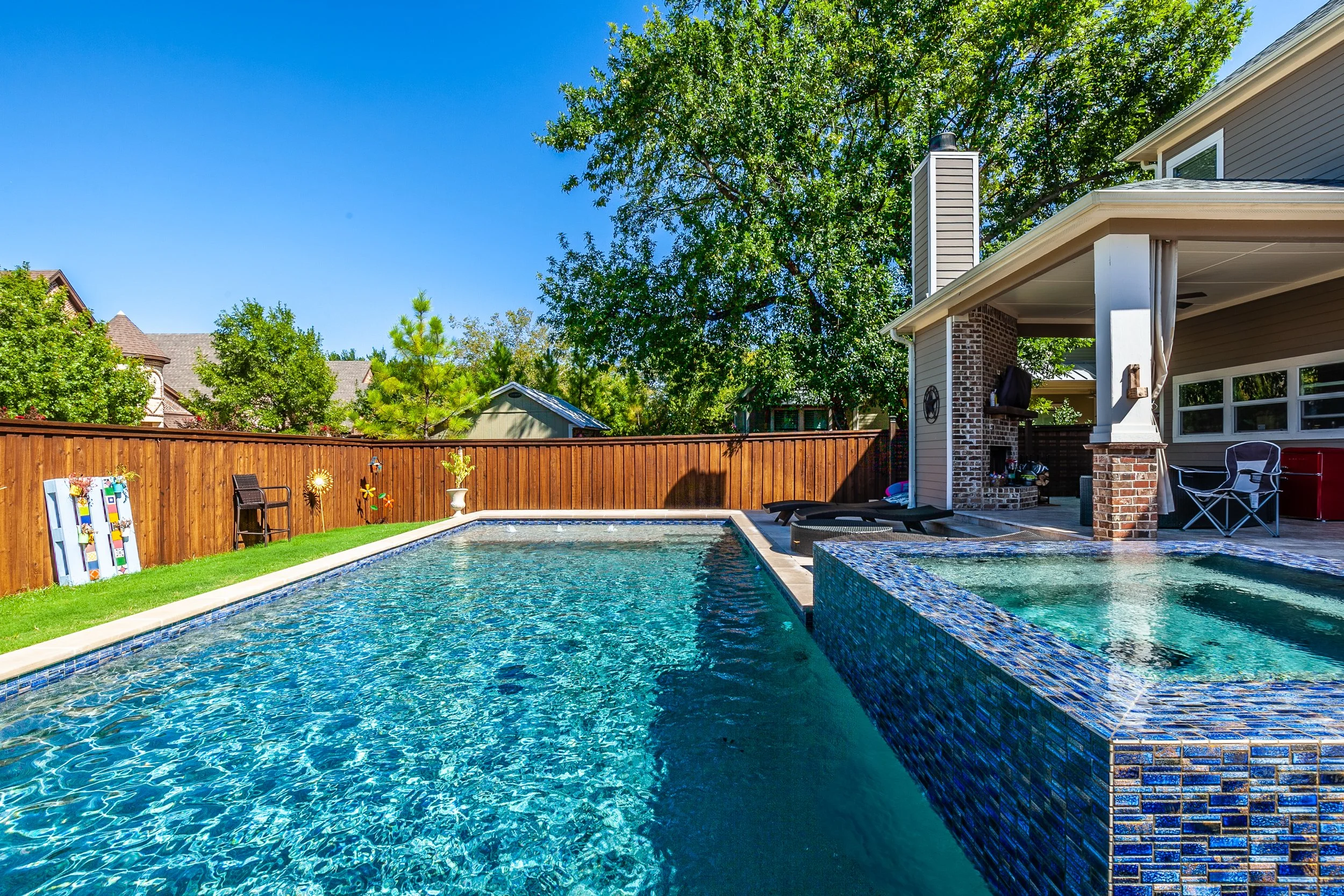 A backyard with a swimming pool, hot tub, and patio area, enclosed by a wooden fence, with trees and neighboring houses in the background.