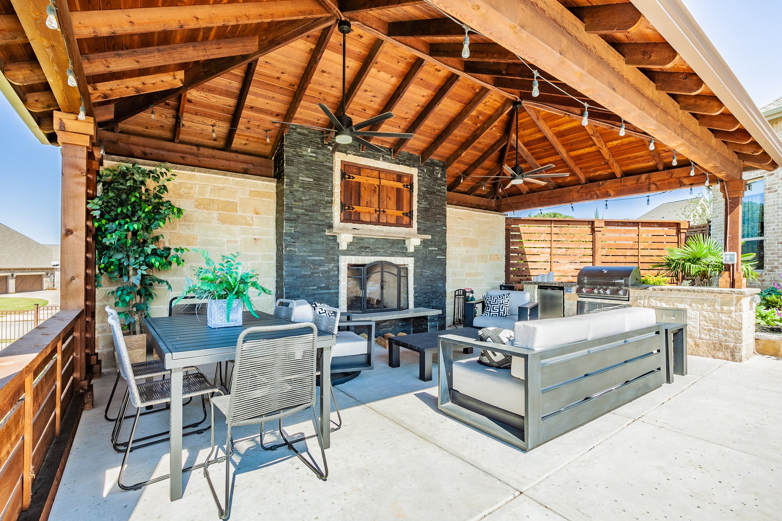 Covered outdoor patio with seating area, fireplace, and grill, decorated with string lights and potted plants.