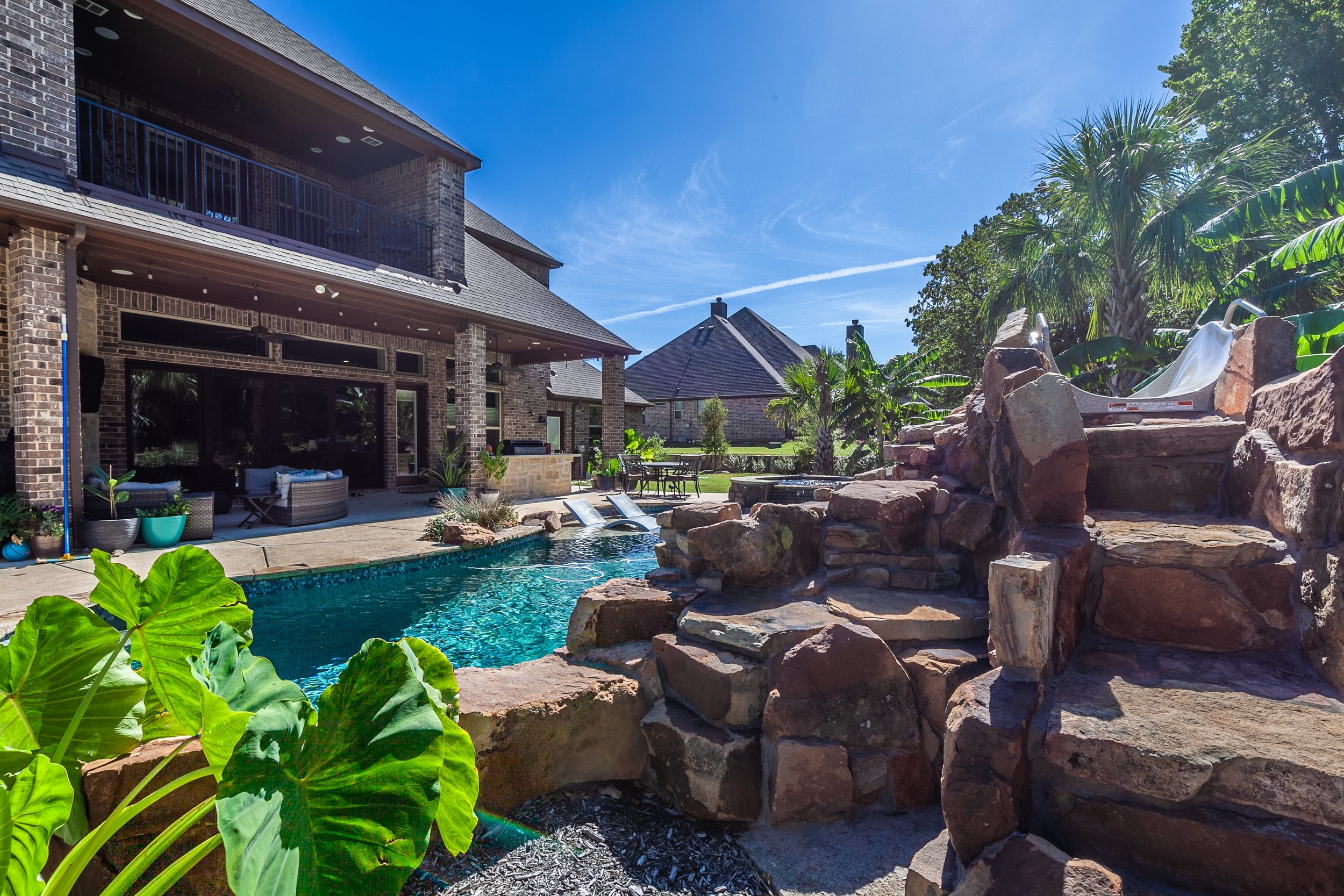 Backyard with swimming pool, rock waterfall feature, outdoor seating area, palm trees, and a slide on rocks, with a brick house and blue sky in the background