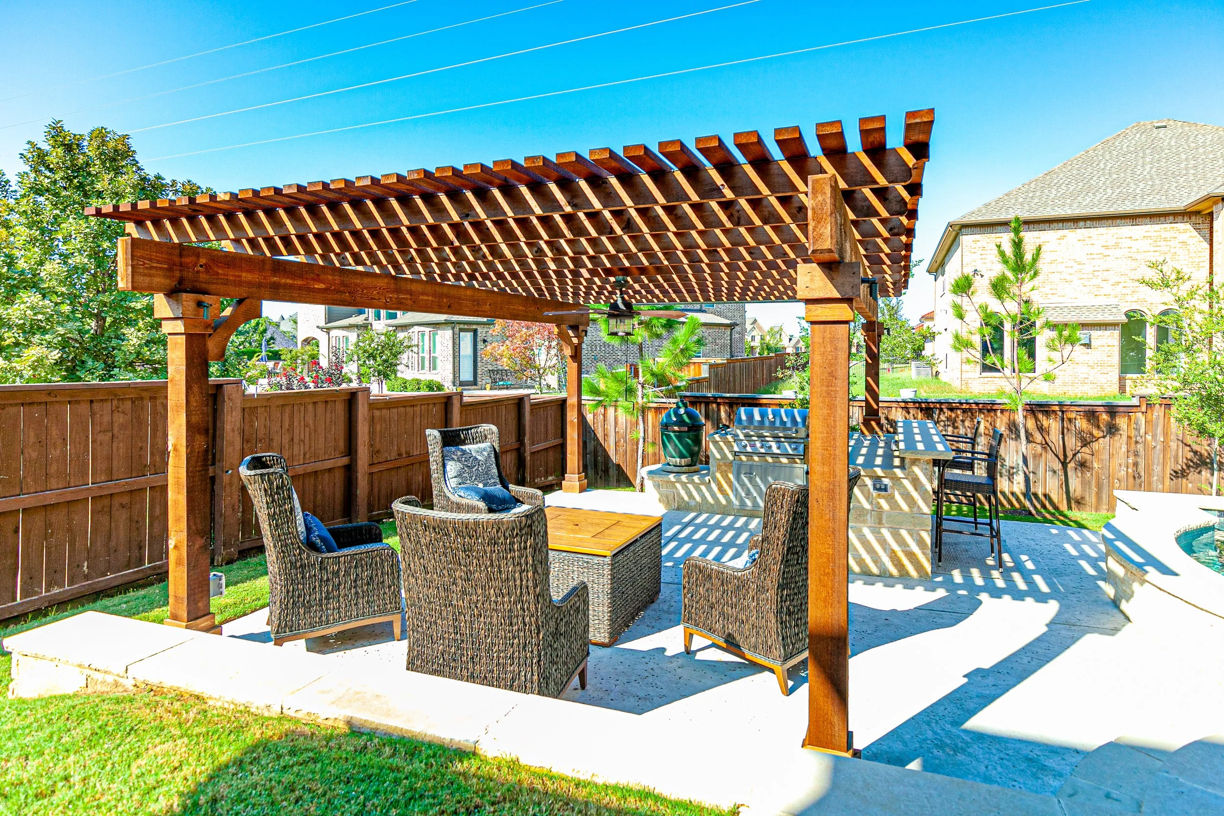 Backyard patio with wooden pergola, wicker chairs around a small table, additional seating area, and nearby trees, fence, and neighbor houses under a clear blue sky.