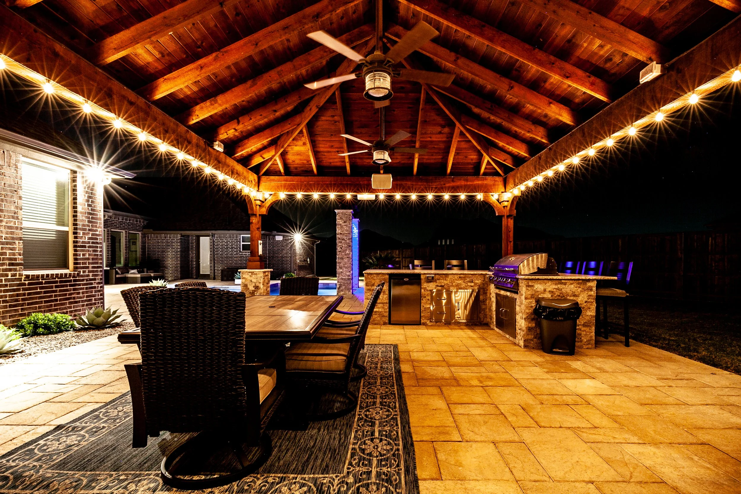 Nighttime outdoor patio with a wooden pergola decorated with string lights, a dining table with chairs, a built-in grill, a small cooler, and outdoor furniture, near a brick house with lit windows.