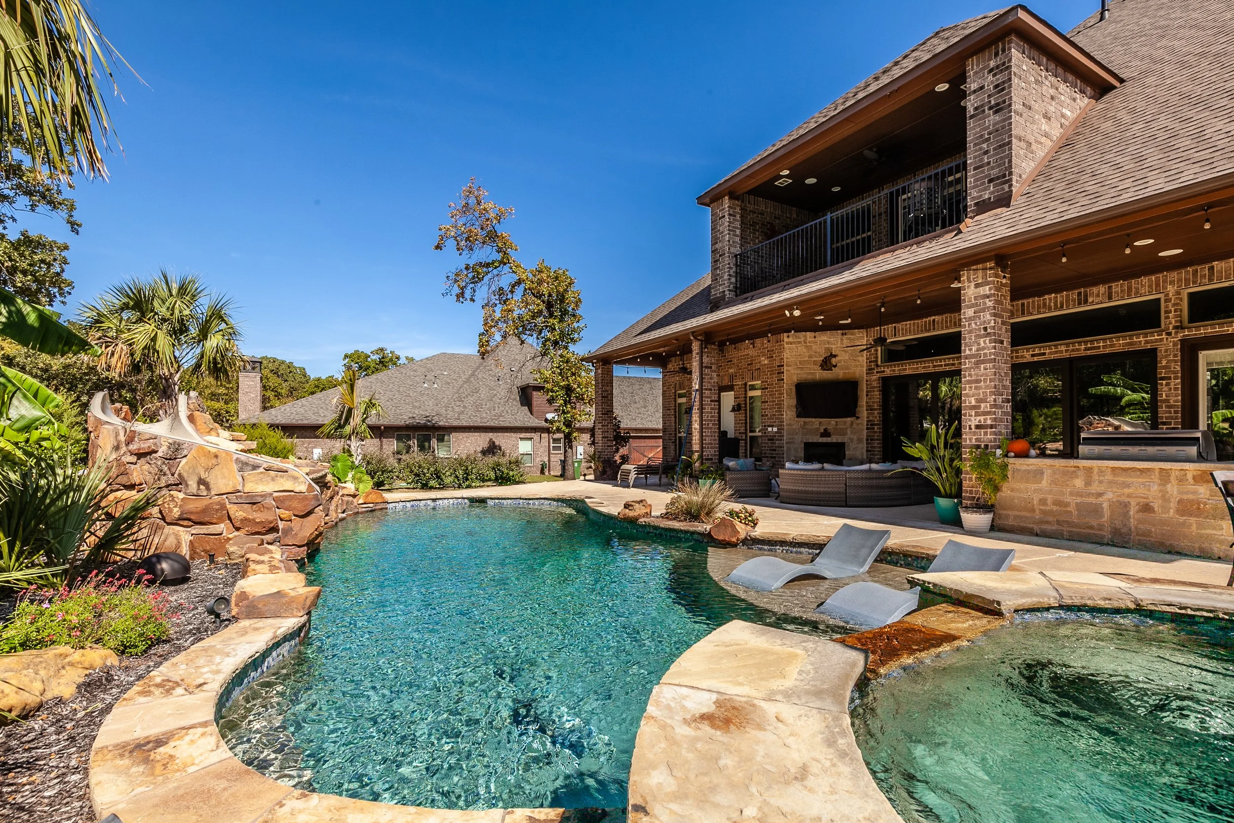 Residential backyard with a kidney-shaped swimming pool, outdoor seating area under a covered patio, and surrounding lush greenery and palm trees.