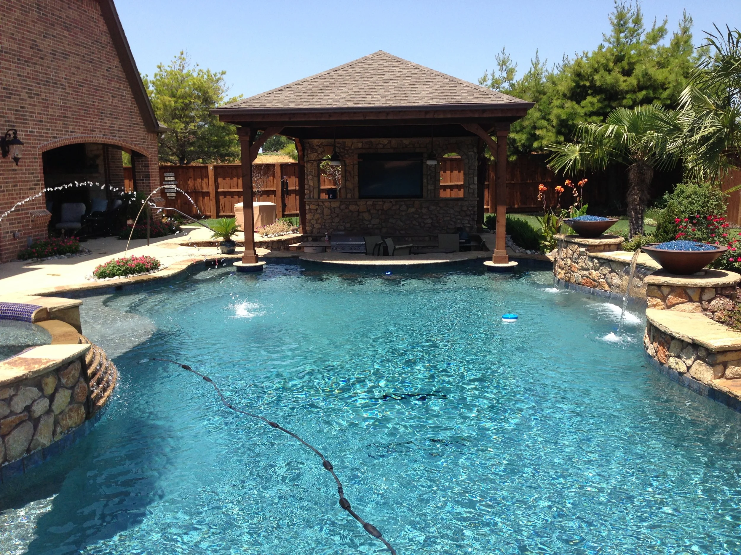 Backyard swimming pool with stone waterfall features, surrounded by landscaped plants and a wooden privacy fence, with a covered patio area in the background.