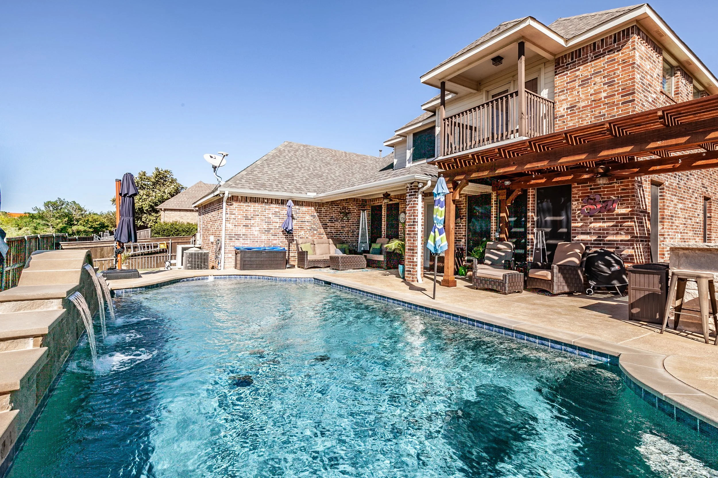 Backyard with a swimming pool, patio furniture, umbrellas, and a two-story brick house with a balcony and pergola under sunny sky.