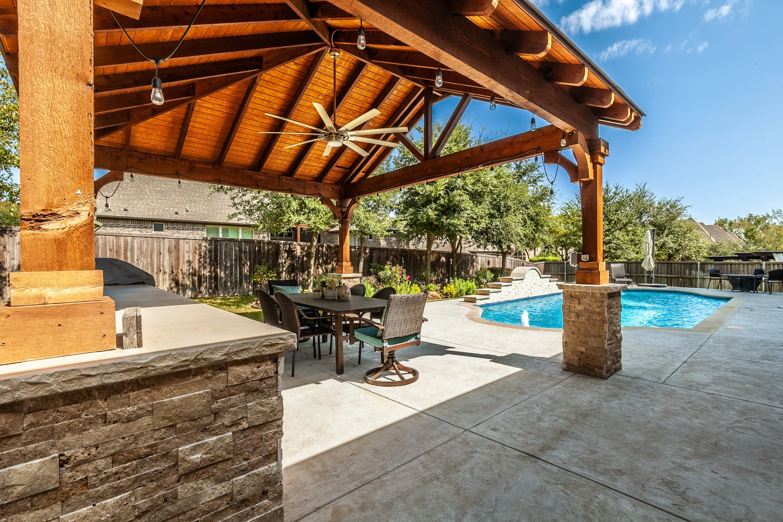Backyard with a covered patio, wooden ceiling, ceiling fan, string lights, dining table with chairs, swimming pool, and fenced yard with trees.