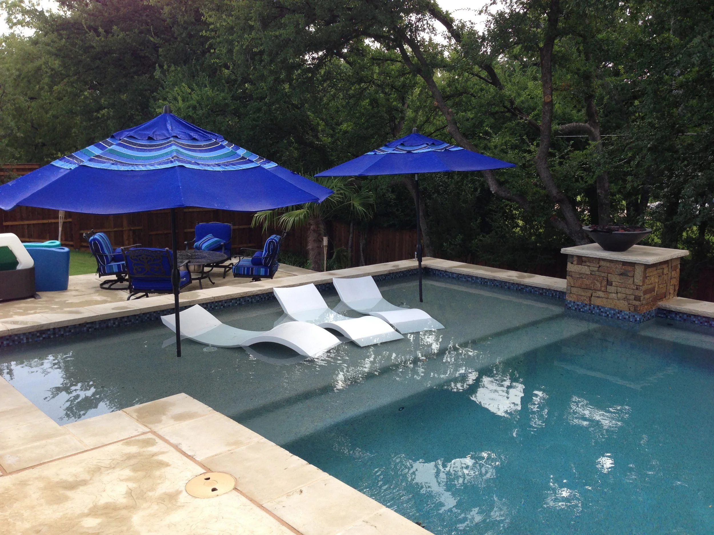 A backyard swimming pool area with two white lounge chairs in the shallow part of the pool, shaded by two blue umbrellas. There is patio furniture with blue cushions and a table with chairs, all shaded by a large blue umbrella. The background has tre