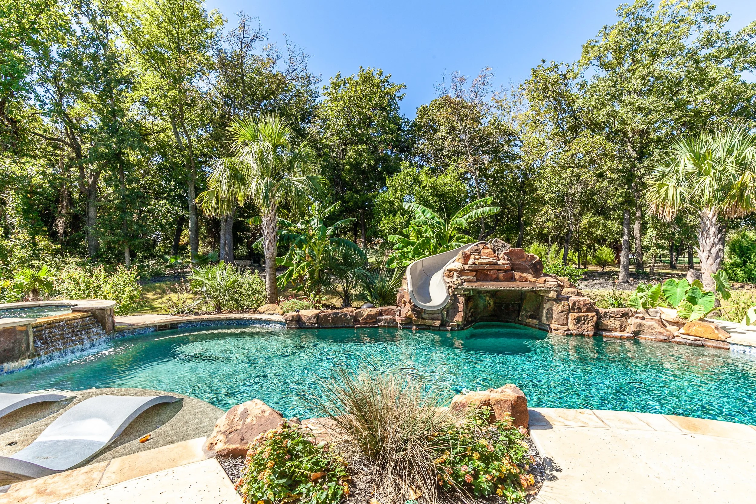 Residential backyard pool with a waterslide, surrounded by lush trees and tropical plants, on a clear sunny day.